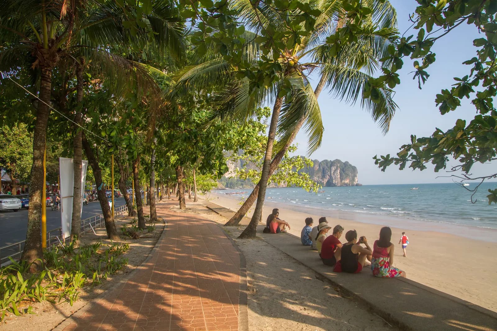 The tree-lined beachfront walk at Ao Nang, with the beach road and parked cars on one side, people sitting on the sea wall and limestone cliffs across the bay