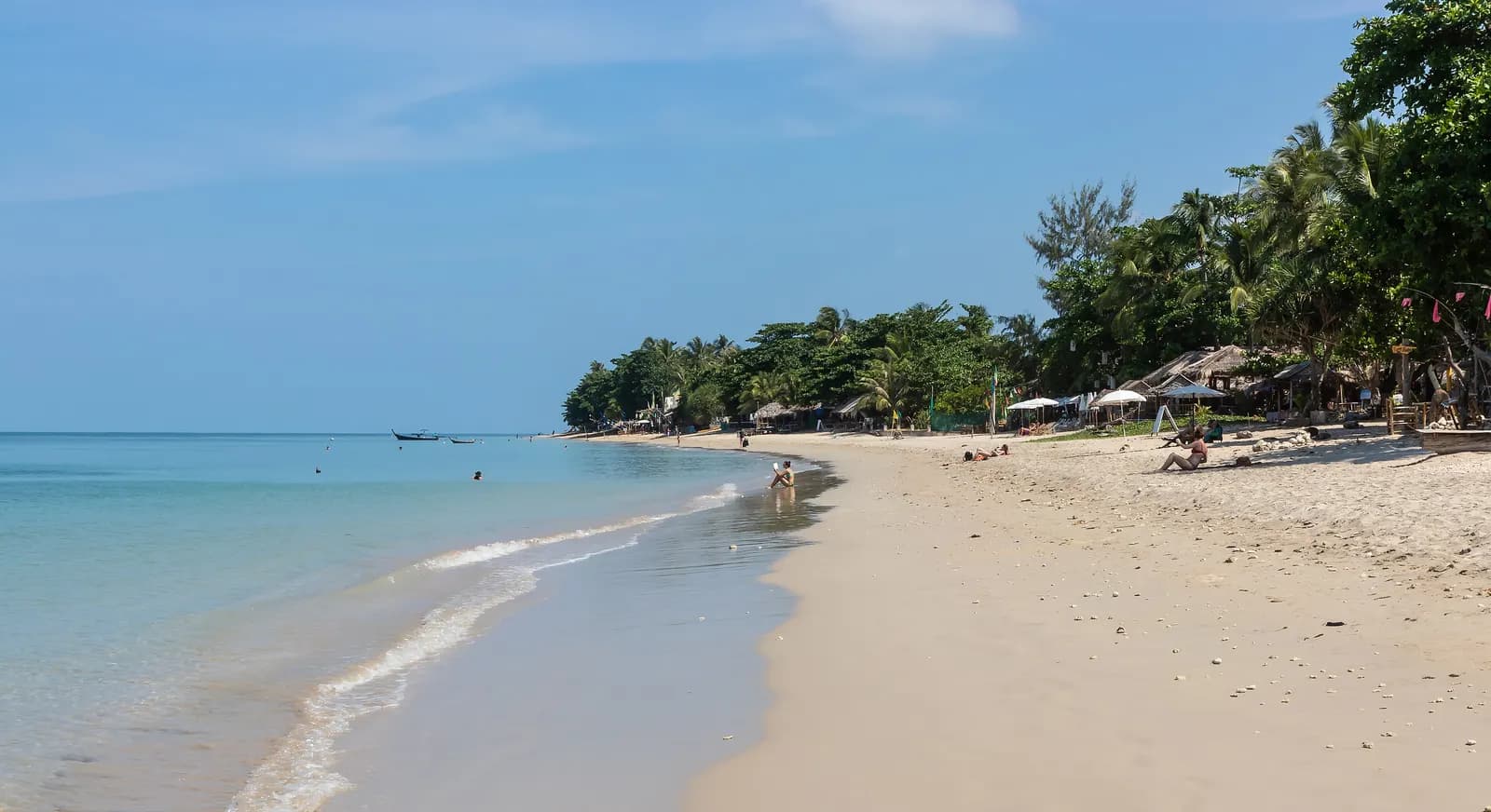 Klong Khong Beach on Ko Lanta, with clear shallow water, longtail boats offshore and thatched beach huts and umbrellas under palm trees