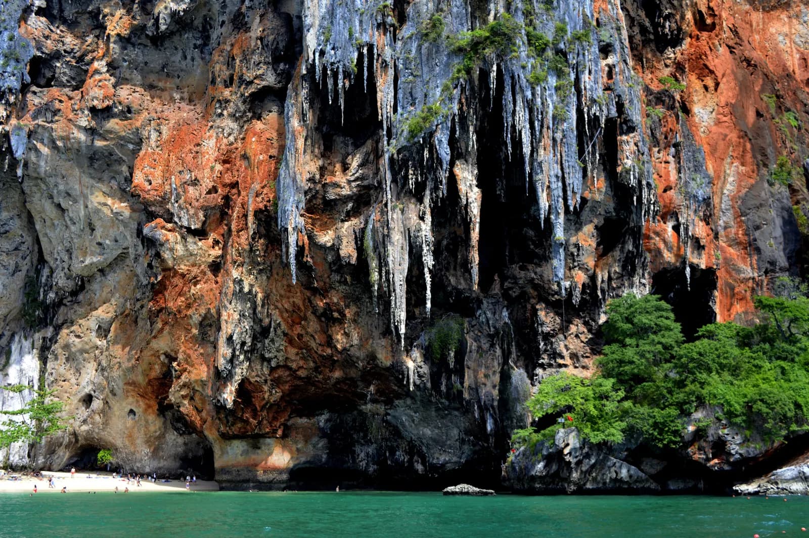 Orange and grey limestone cliffs with hanging rock formations above a small beach and green water on the Railay peninsula