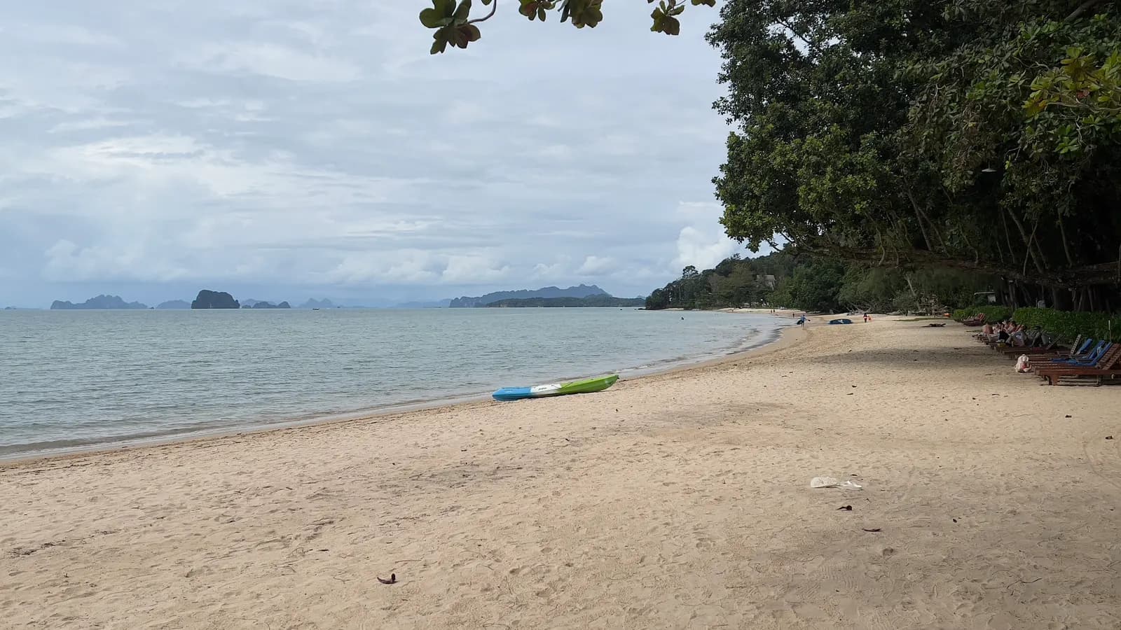 Tubkaek Beach under a cloudy sky, with a kayak on the sand, sun loungers under the trees and limestone islands on the horizon