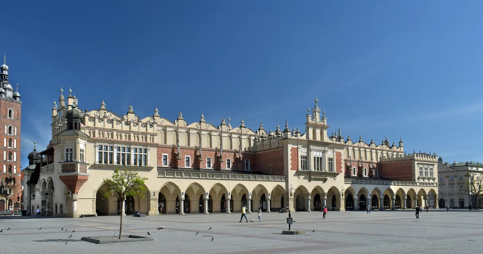 The long arcaded Cloth Hall in the middle of Kraków's Main Market Square under a blue sky, with a church tower at the left edge