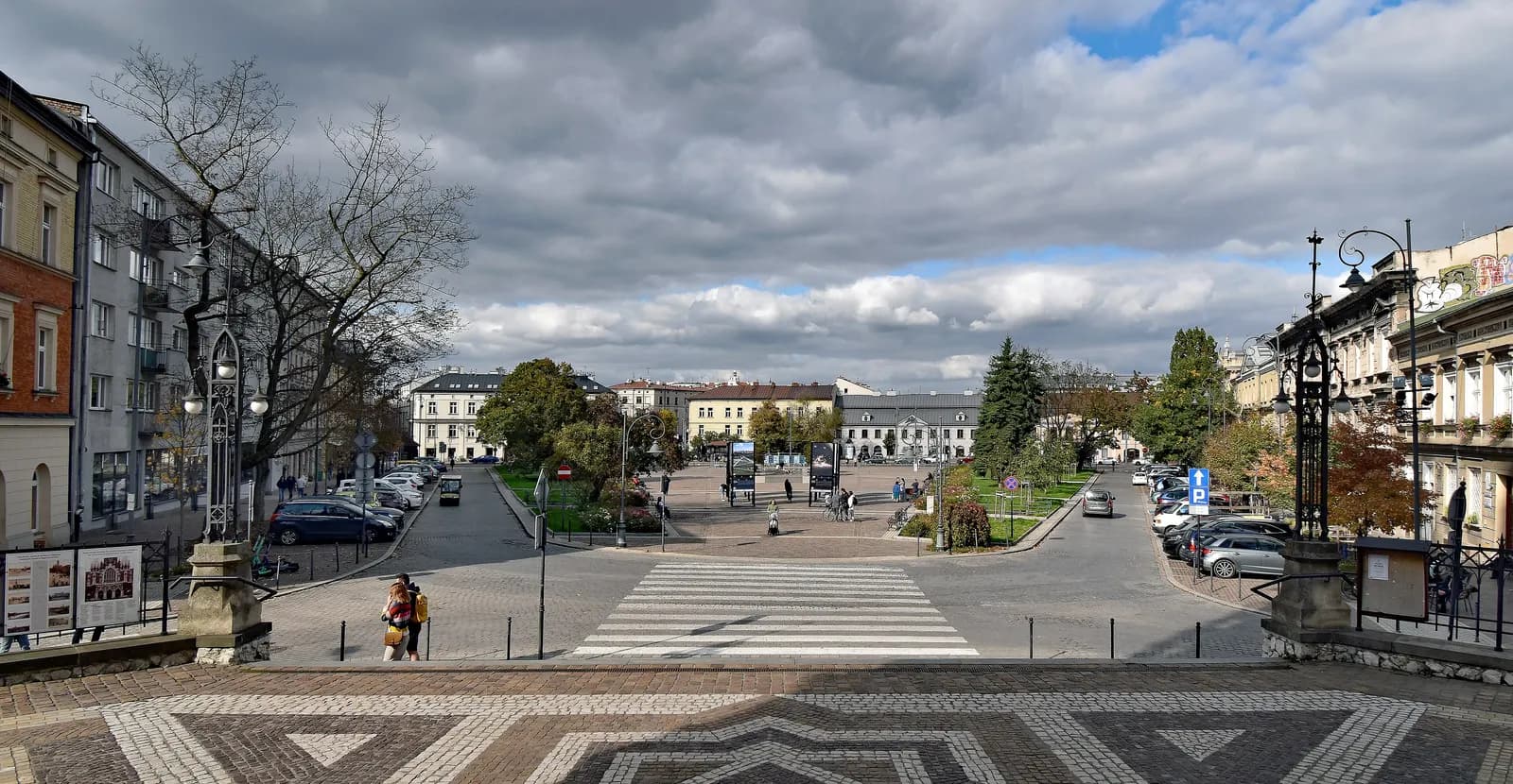Podgórski Market Square in Kraków's Podgórze district seen from its southern end, with a patterned pavement, a pedestrian crossing, trees and townhouses around the square