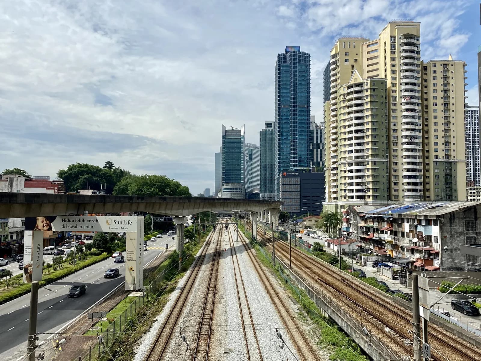 Railway tracks running through Brickfields towards the towers around KL Sentral, with an elevated rail line crossing overhead, a main road on the left and apartment blocks on the right