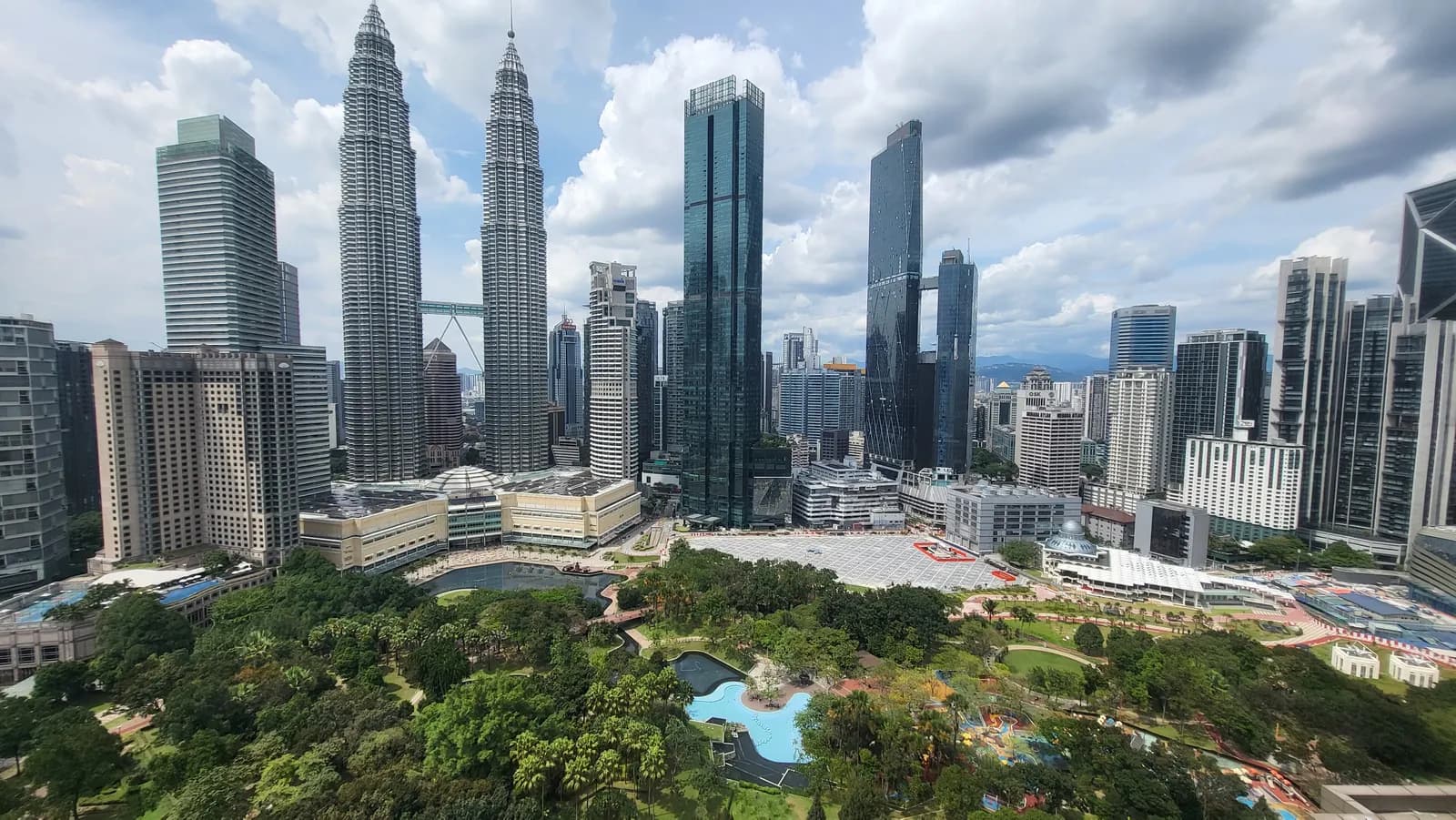 The Petronas Twin Towers and neighbouring office towers above the gardens, lake and wading pool of KLCC Park, seen from high up on a bright, cloudy day