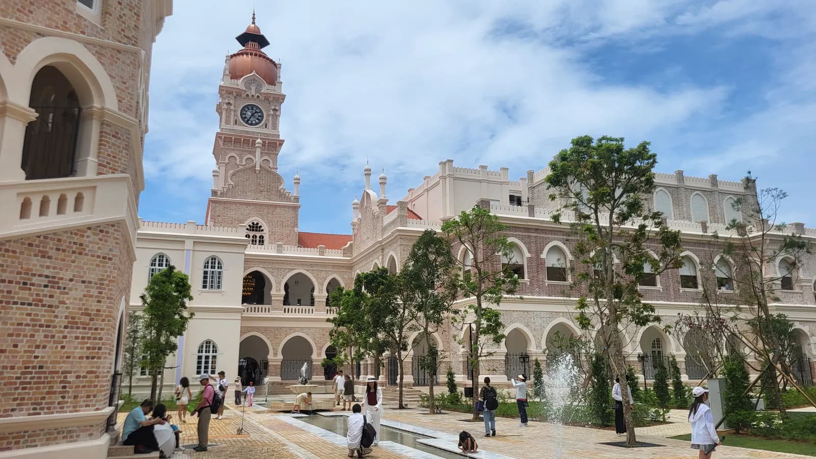 The clock tower and arched brick facades of the Sultan Abdul Samad Building around a courtyard with fountains, young trees and visitors, after its renovation