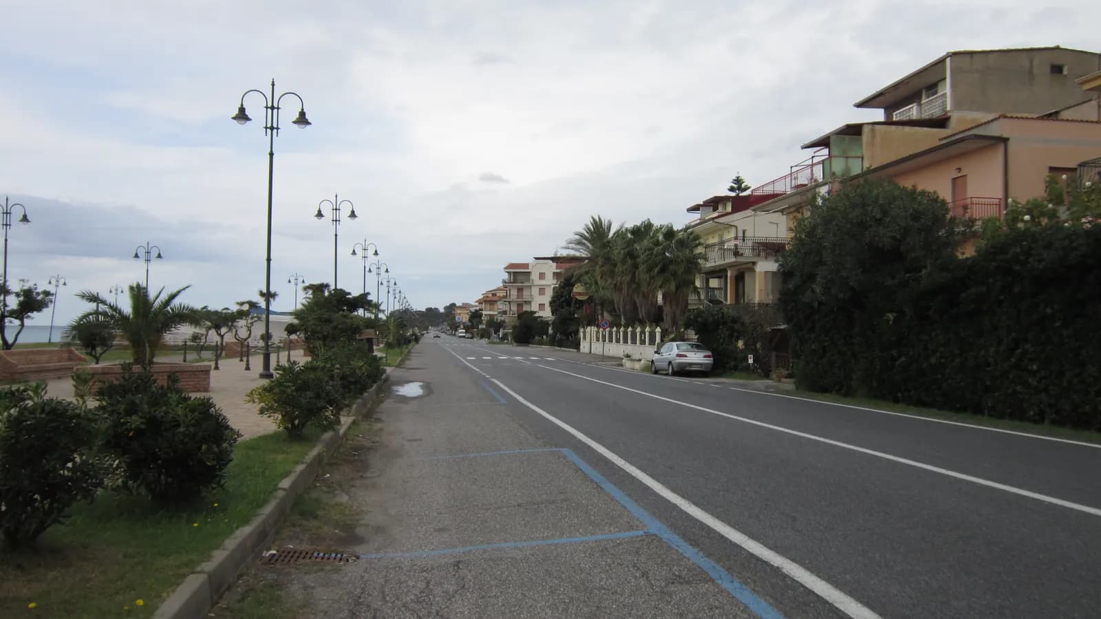 The seafront road at Falerna Lido, with blue-marked parking bays along the kerb, lamp posts and palms on the promenade side and low apartment blocks on the other.