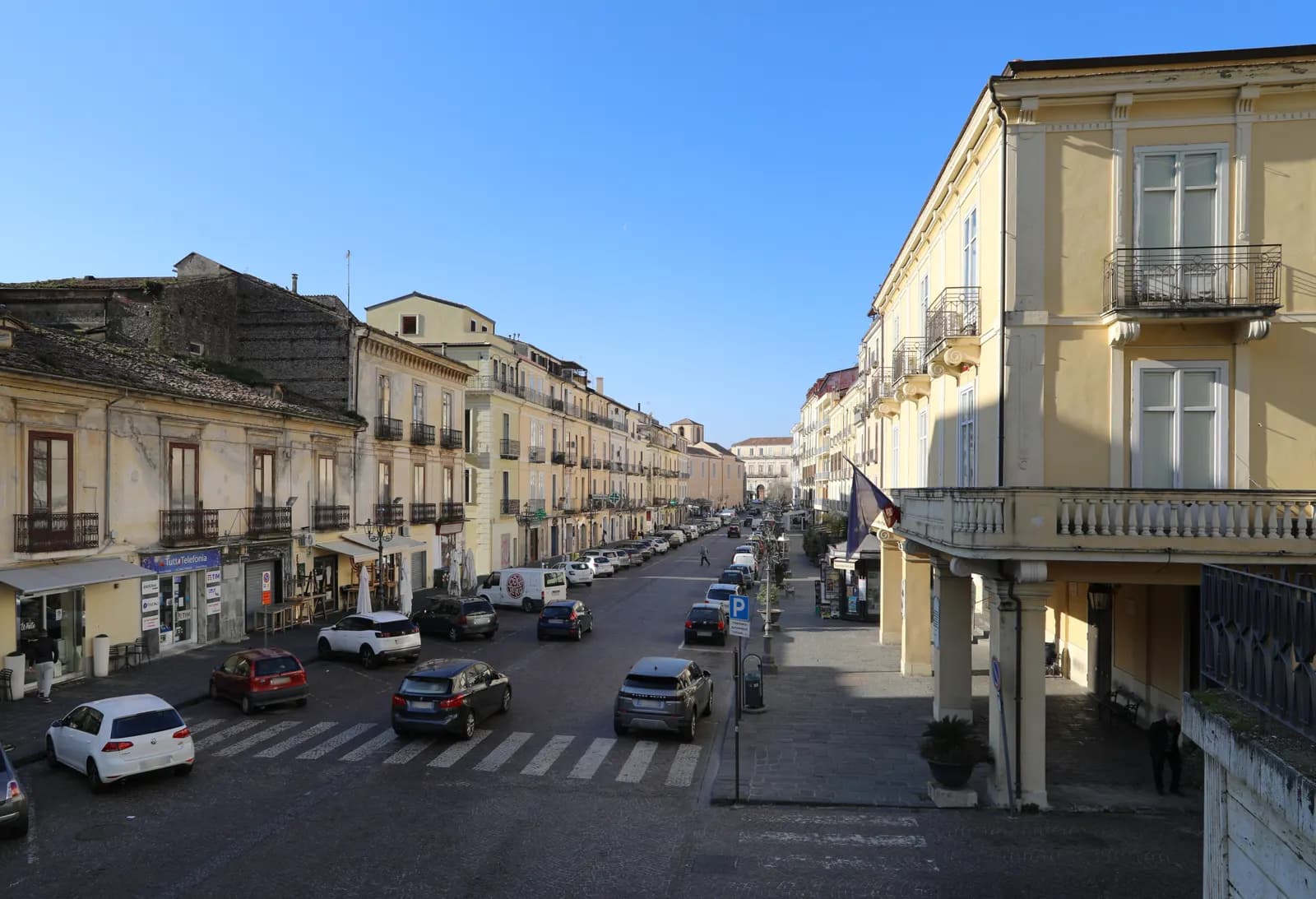 Corso Numistrano in Nicastro, the main street of Lamezia Terme, with cars parked along both kerbs, a blue parking sign and tall pastel buildings with balconies on each side.