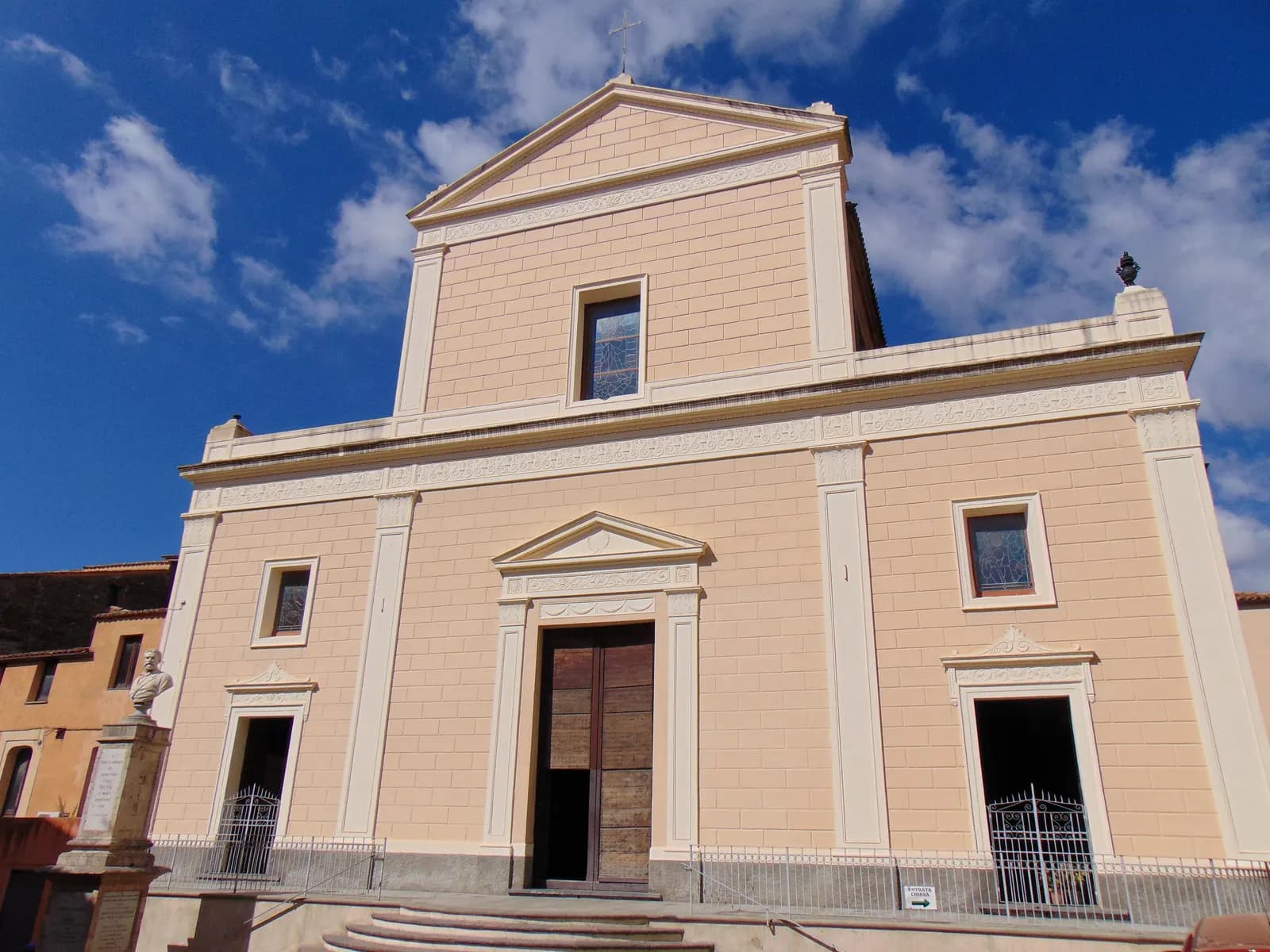 The plain cream front of the Chiesa Matrice in Sambiase, with pilasters, a triangular pediment and a railed terrace, under a blue sky with scattered cloud.