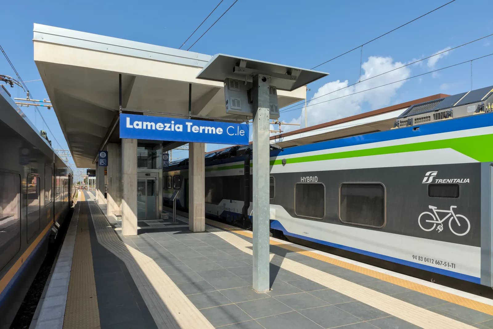 A platform at Lamezia Terme Centrale station under a blue sky, with the station name on a blue sign and a green and grey Trenitalia regional train alongside.