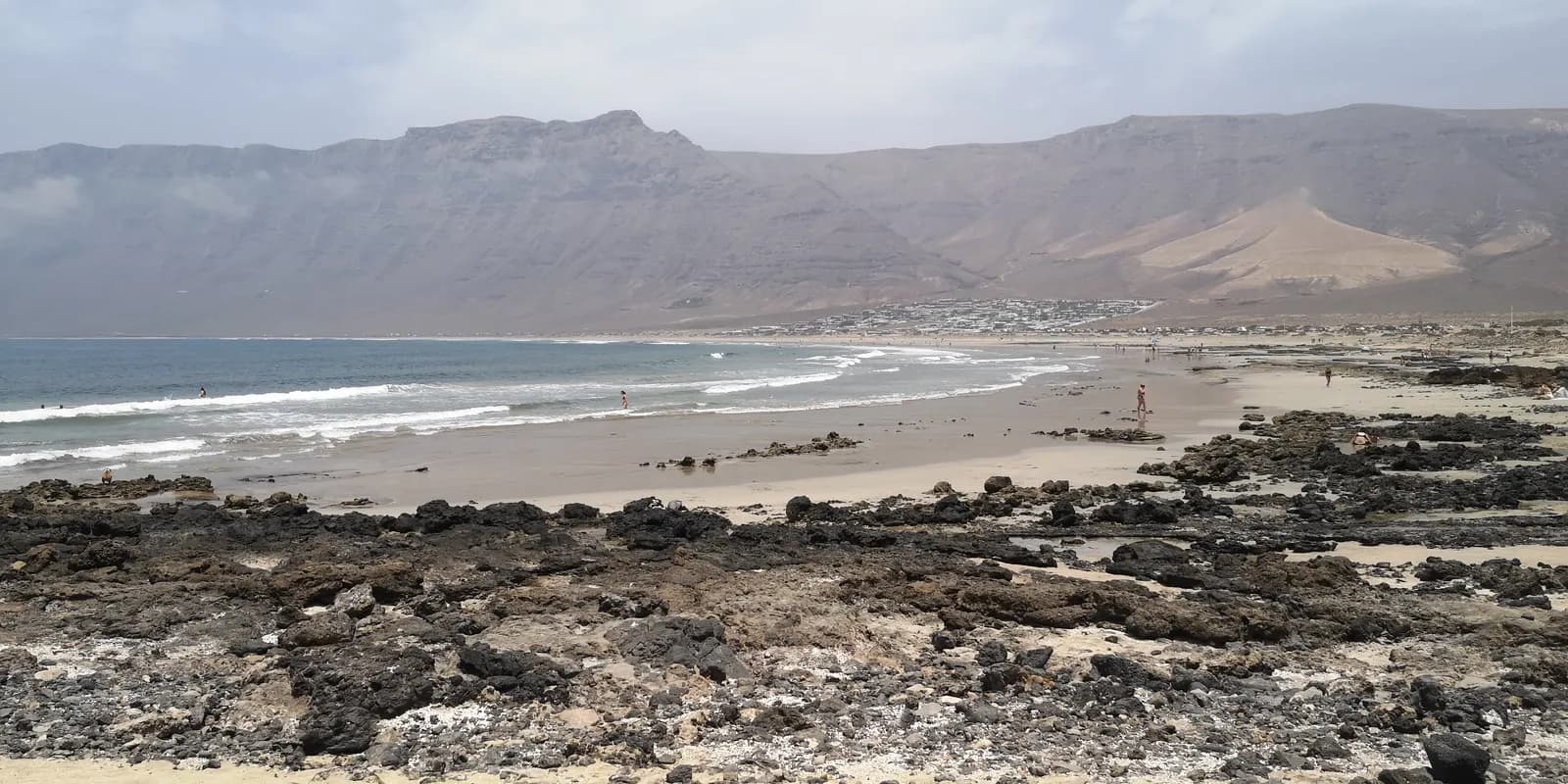 The beach at Caleta de Famara with black volcanic rocks and waves, below the long cliffs of the Risco de Famara.