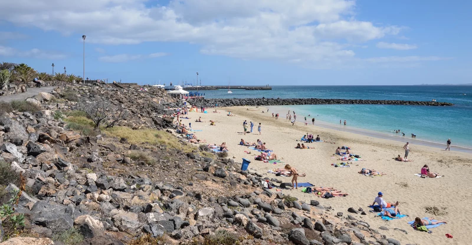 The sandy town beach of Playa Blanca with sunbathers, seen from the rocky slope above it, looking towards the breakwater and the marina.