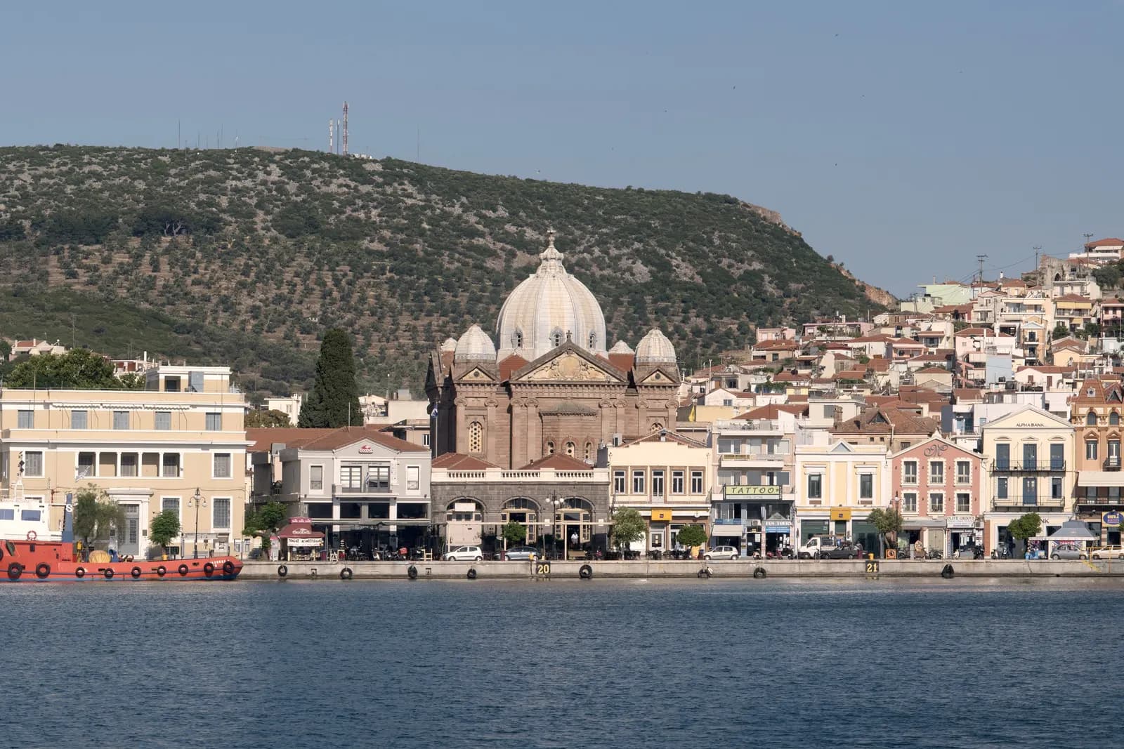 The Mytilene waterfront with a large domed church, cafes and houses below a wooded hill, seen across the harbour