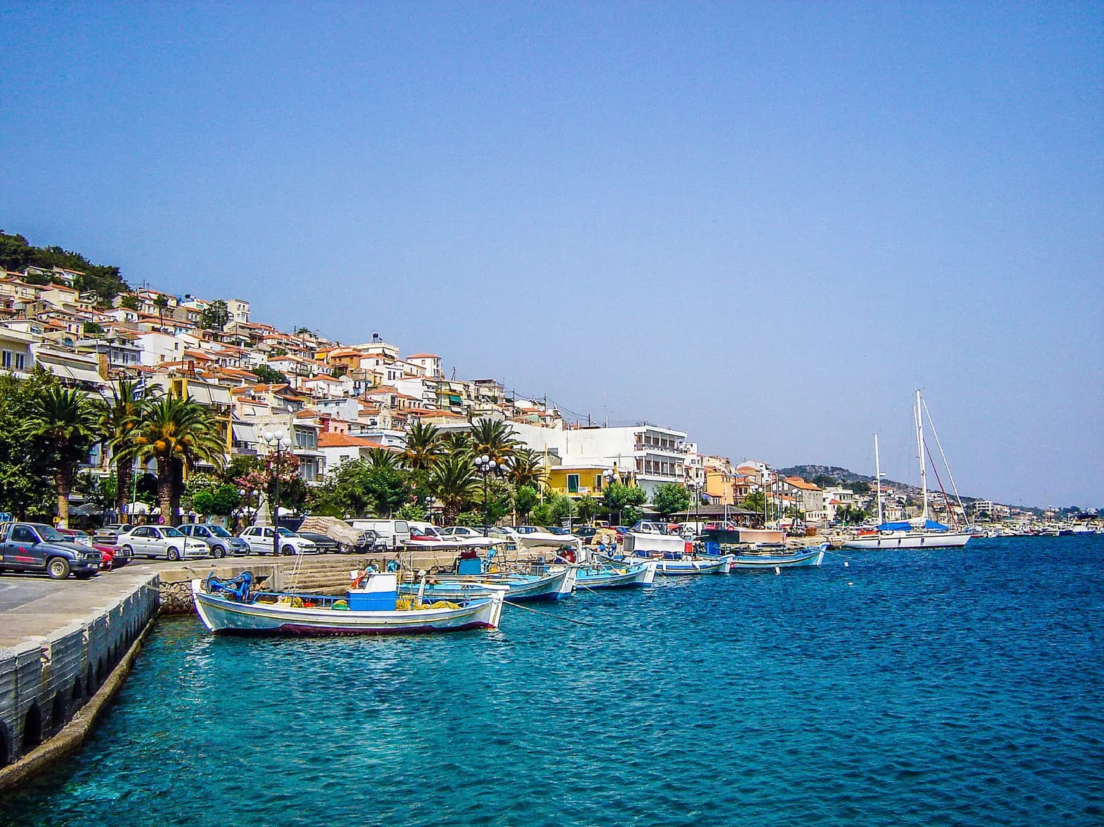 The seafront of Plomari with fishing boats, palm trees, parked cars and houses on the hillside