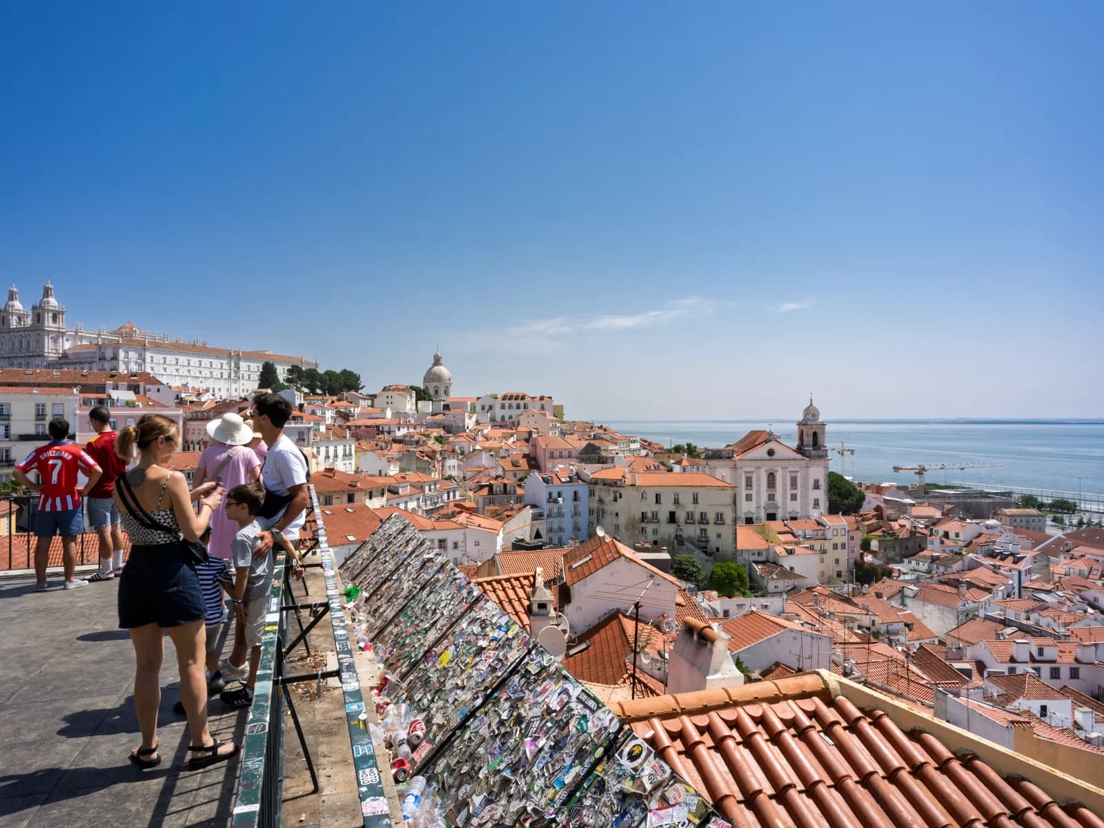 Visitors at the Miradouro de Santa Luzia looking over the terracotta rooftops of Alfama towards the Tagus on a clear day.