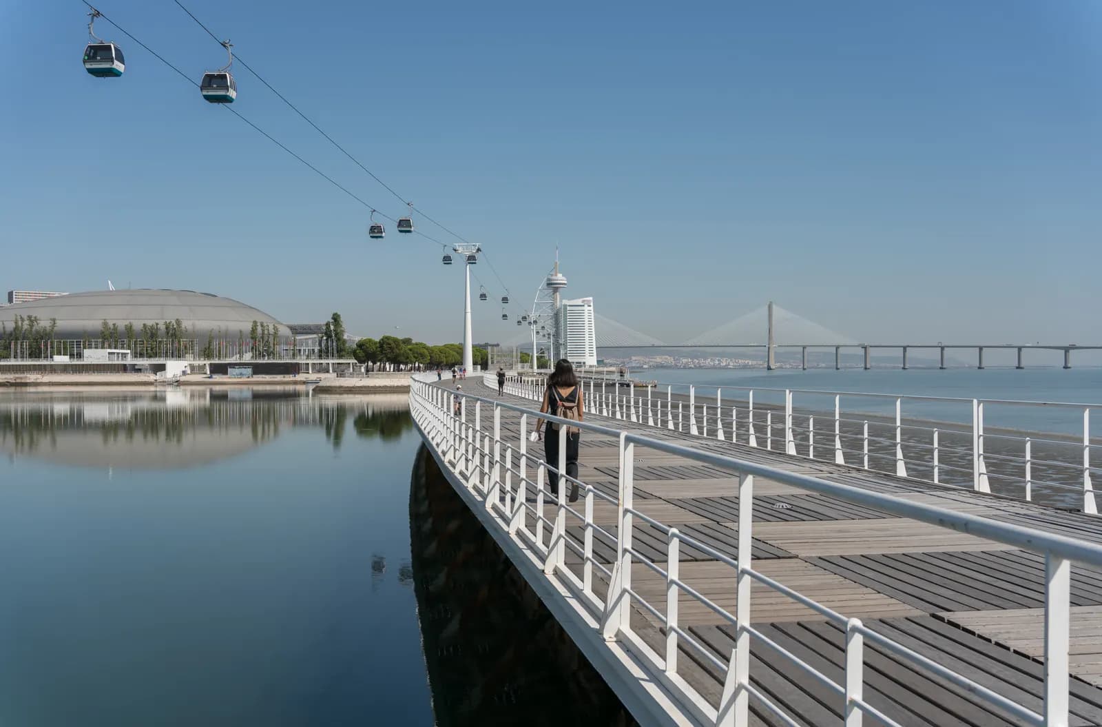 Riverside boardwalk in Parque das Nações with the cable car overhead and the Vasco da Gama Bridge on the horizon.