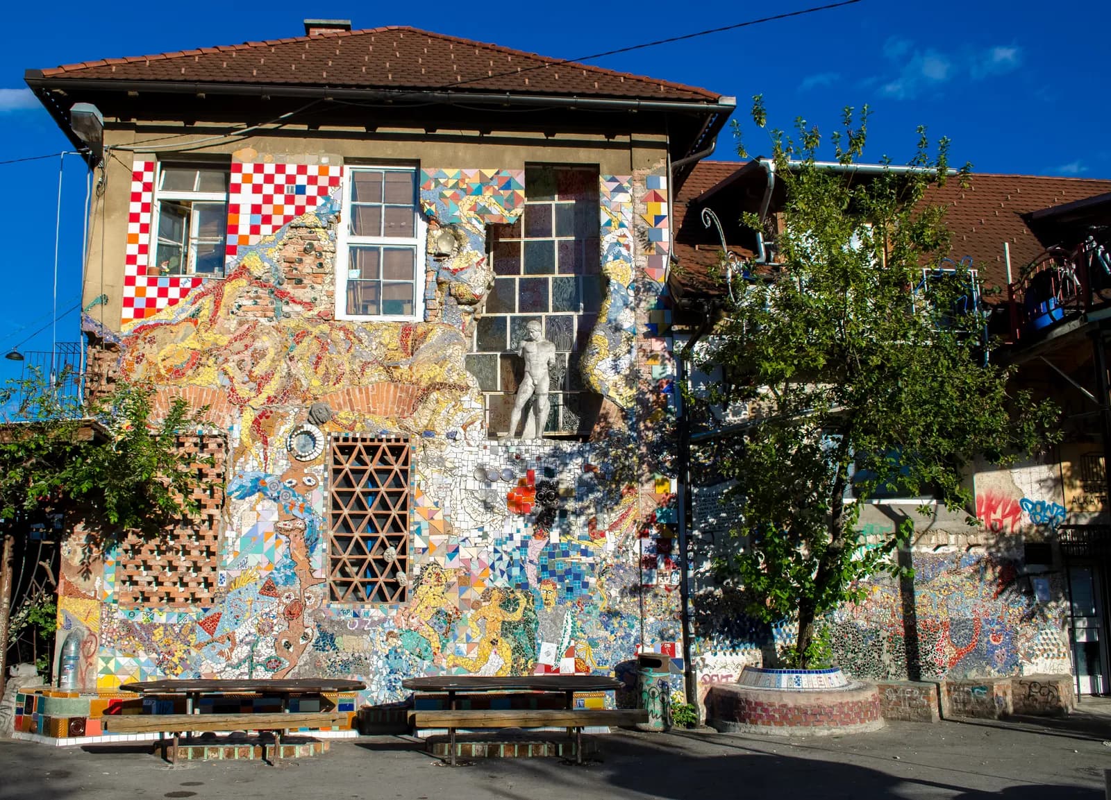 A building at Metelkova mesto in Ljubljana covered in mosaics and murals, with a statue on the wall and benches in front