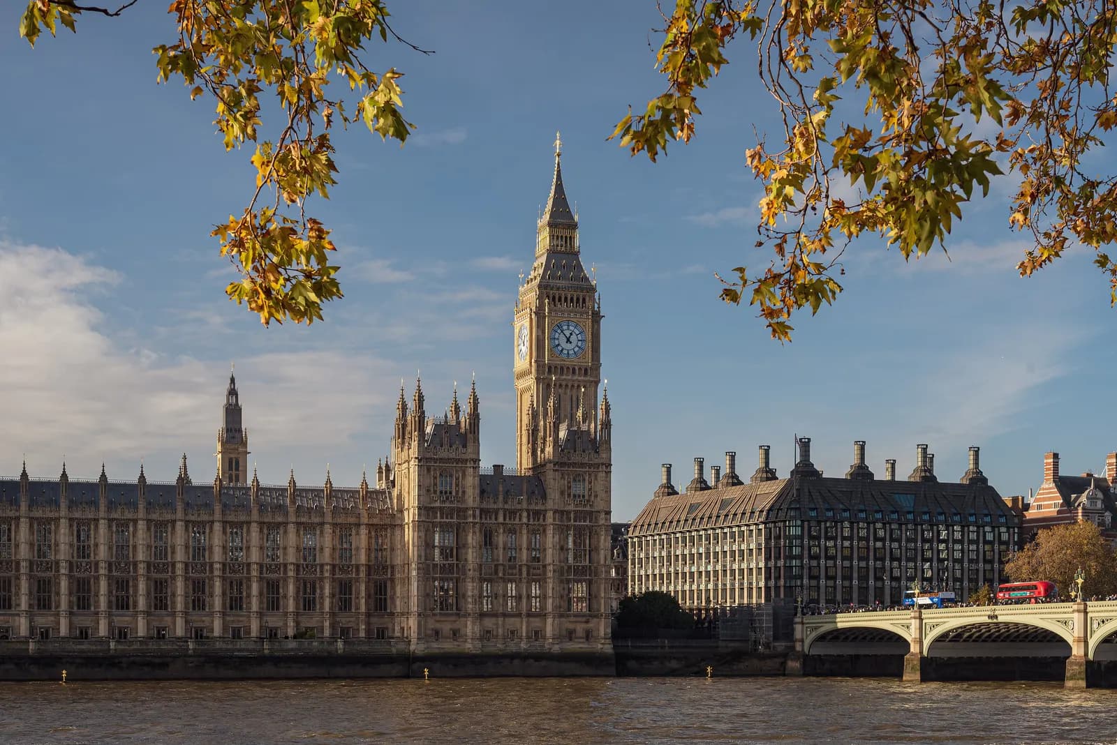 The Palace of Westminster, Elizabeth Tower and Westminster Bridge seen across the Thames from the east bank, framed by autumn leaves
