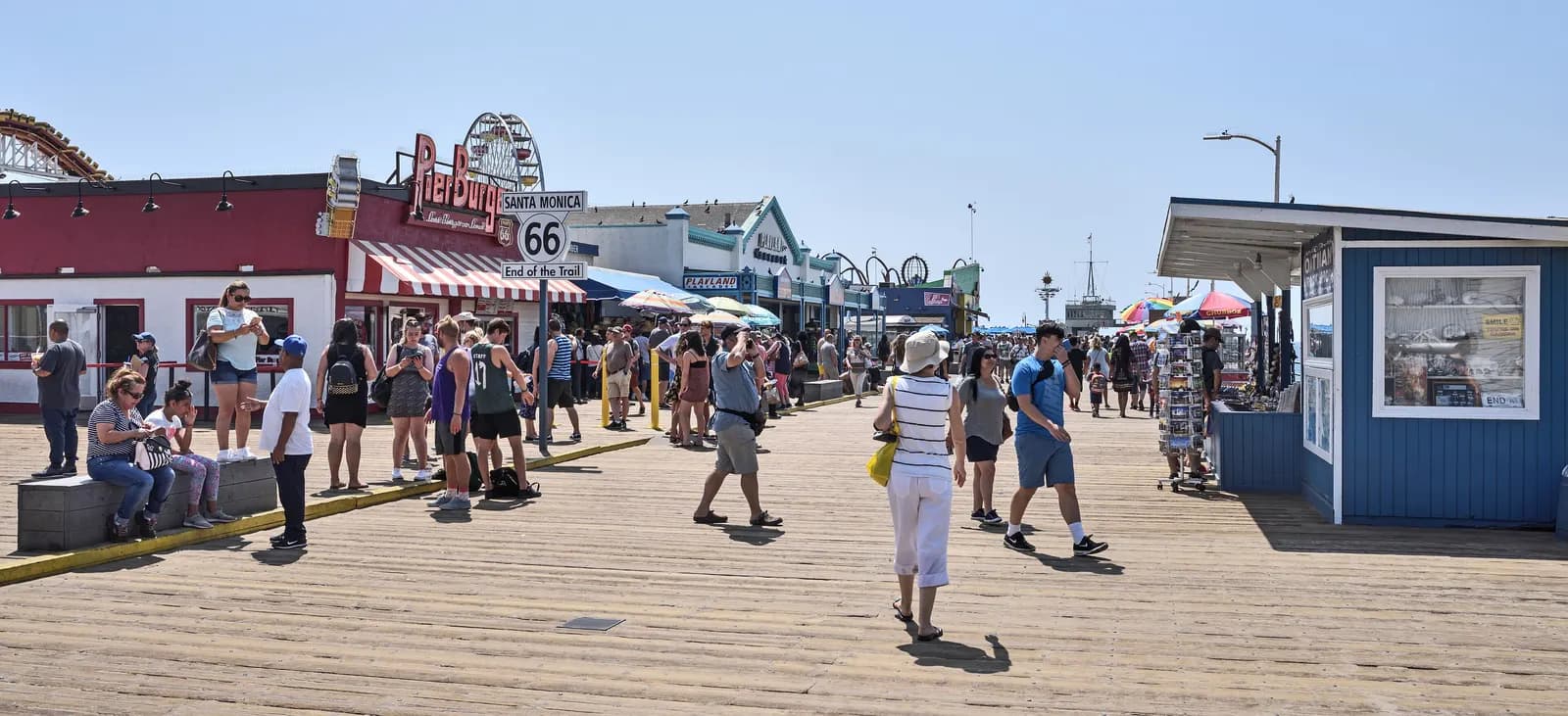 Visitors walking on the wooden deck of Santa Monica Pier past the Route 66 End of the Trail sign, with shops and the Ferris wheel behind
