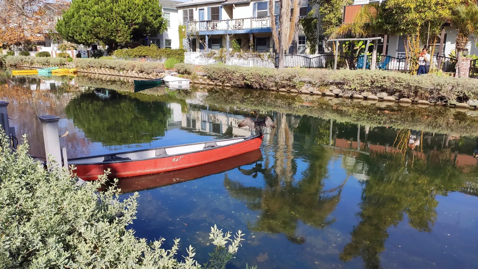 A red canoe moored on one of the Venice canals, with houses, gardens and palm trees reflected in the water