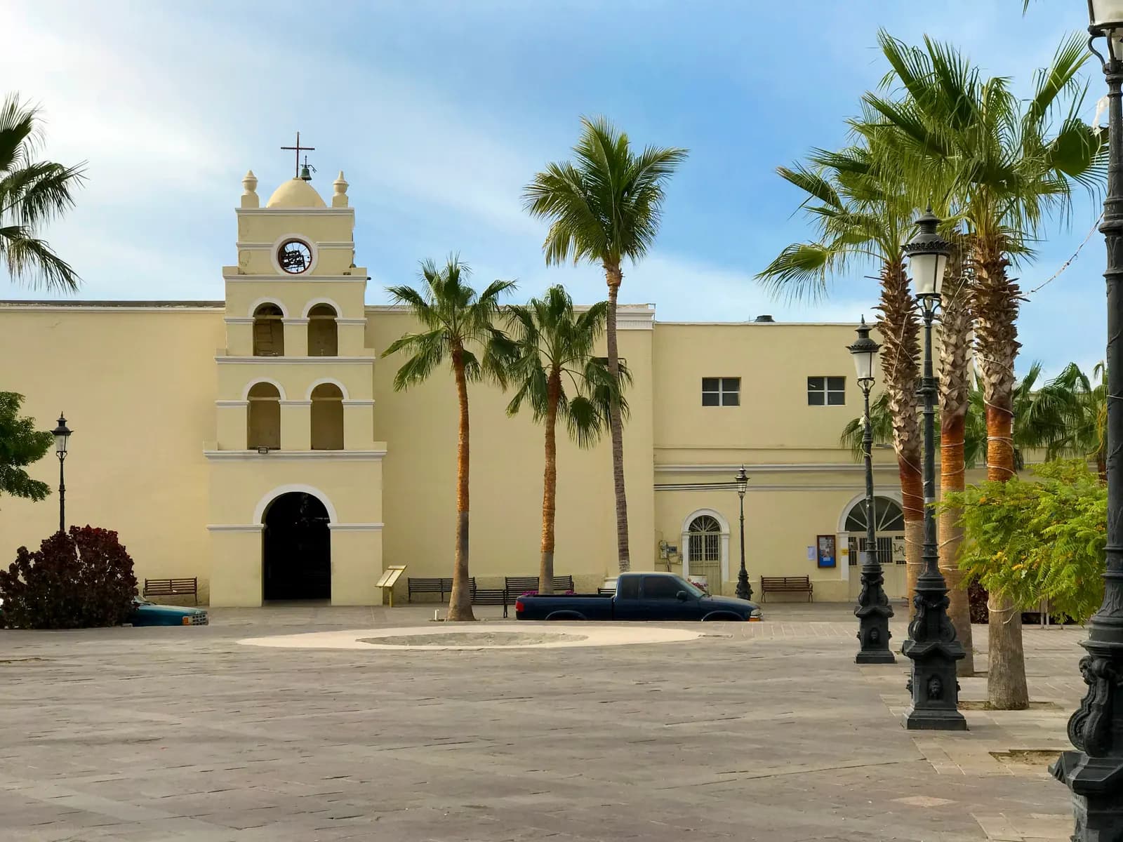 The pale yellow mission church of Todos Santos with its bell tower, palm trees and a paved plaza in front