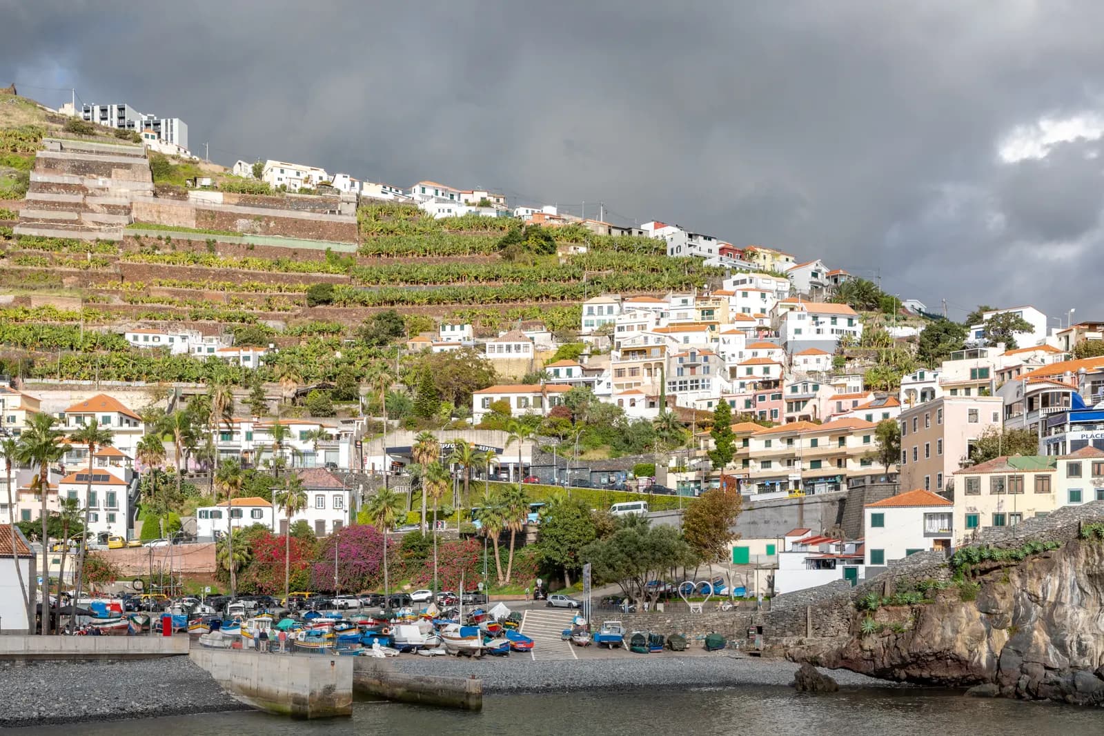 Câmara de Lobos harbour with fishing boats pulled up on the shore and white houses climbing terraced banana plots under a cloudy sky