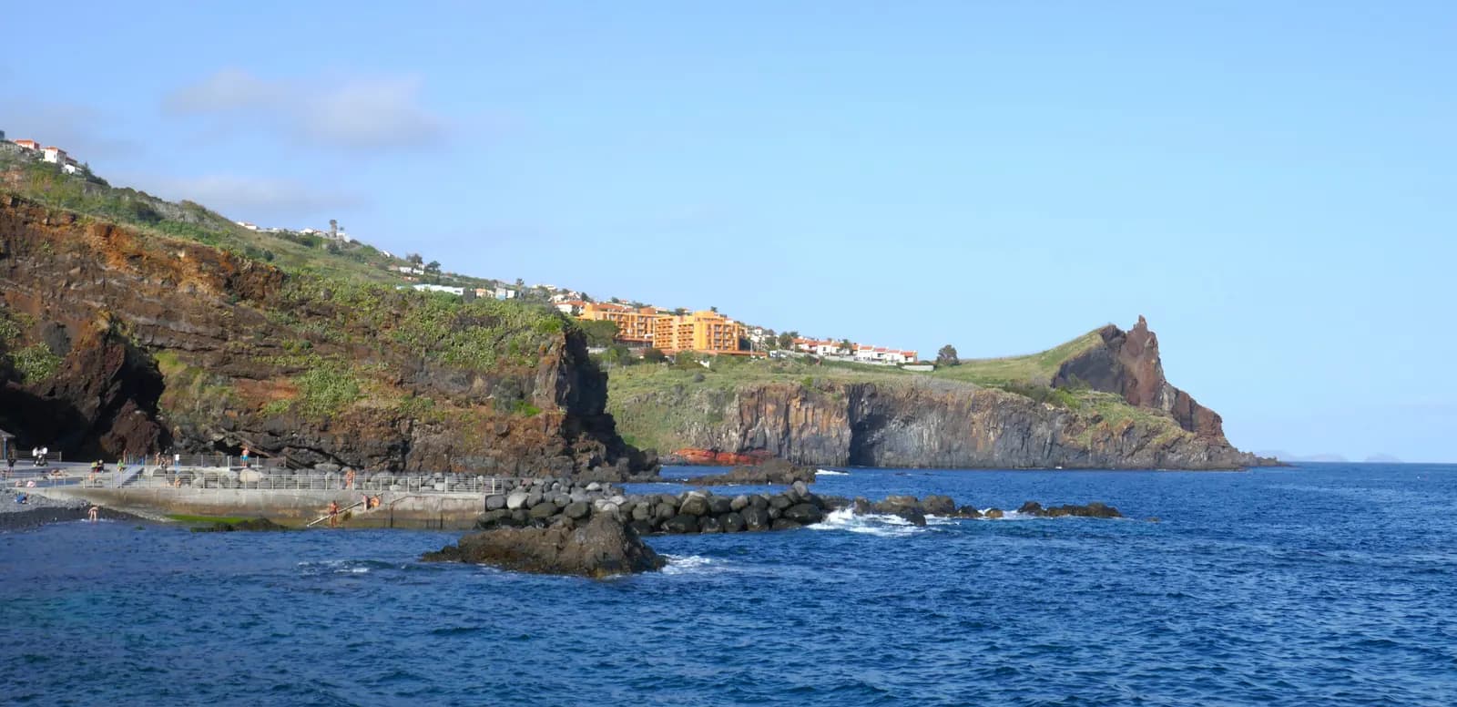 The coast at Reis Magos in Caniço, with dark cliffs, a seafront walkway and buildings on the headland above a blue sea