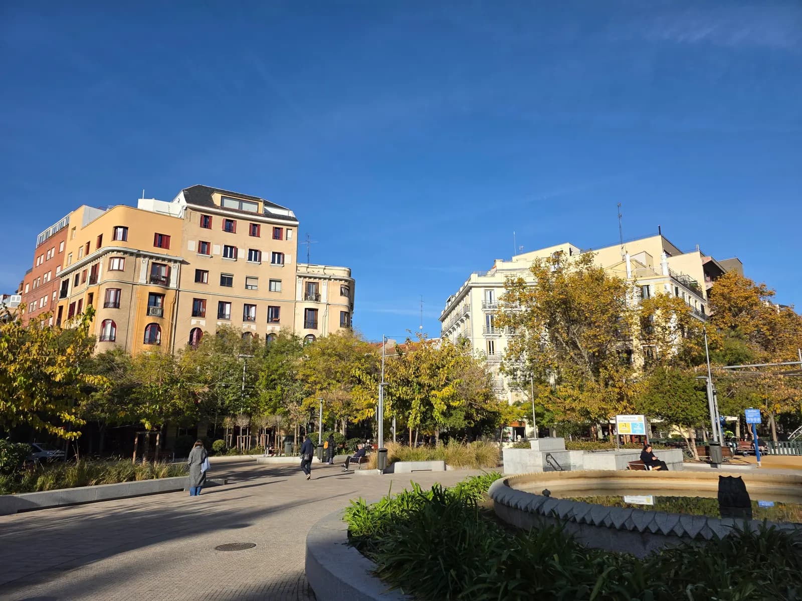 Plaza de Olavide in Chamberí, a round square with trees, benches and a fountain, ringed by residential buildings under a blue sky.