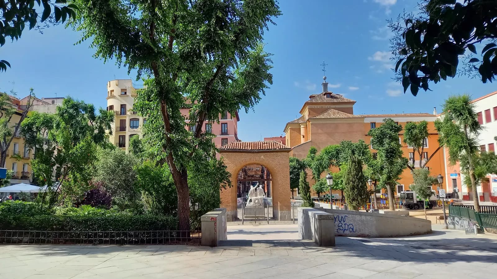 Plaza del Dos de Mayo in Malasaña, a tree-shaded square with a brick arch at its centre and ochre houses around it.
