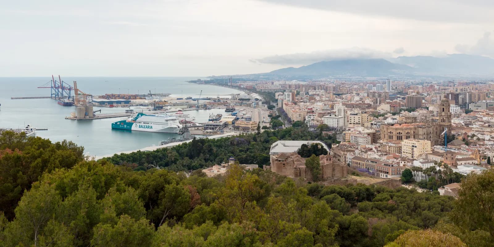 Málaga seen from Gibralfaro: the port with a docked ferry, the cathedral and the old town, and the coast curving west.