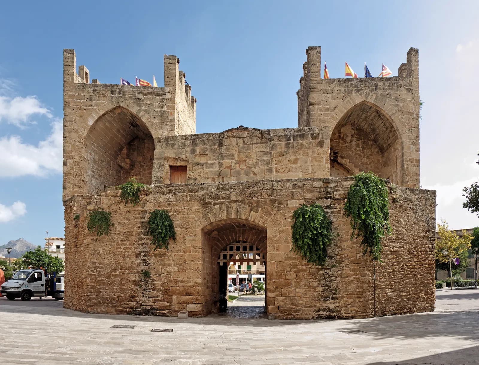 The Porta del Moll, a stone gate with two towers in the town walls of Alcúdia, under a blue sky.