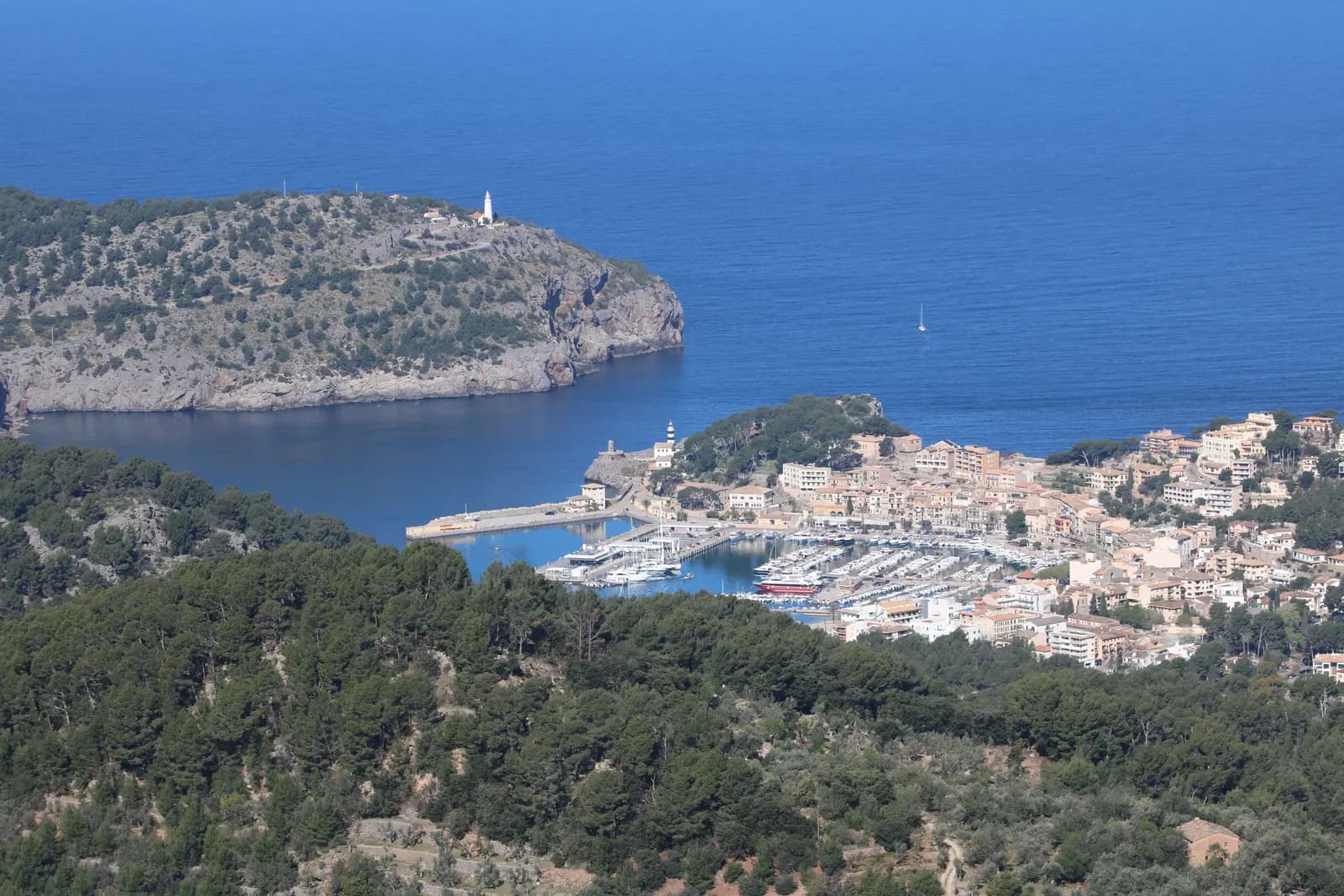 Port de Sóller seen from the hills, with its marina and houses on a round bay closed by a wooded headland with a lighthouse.