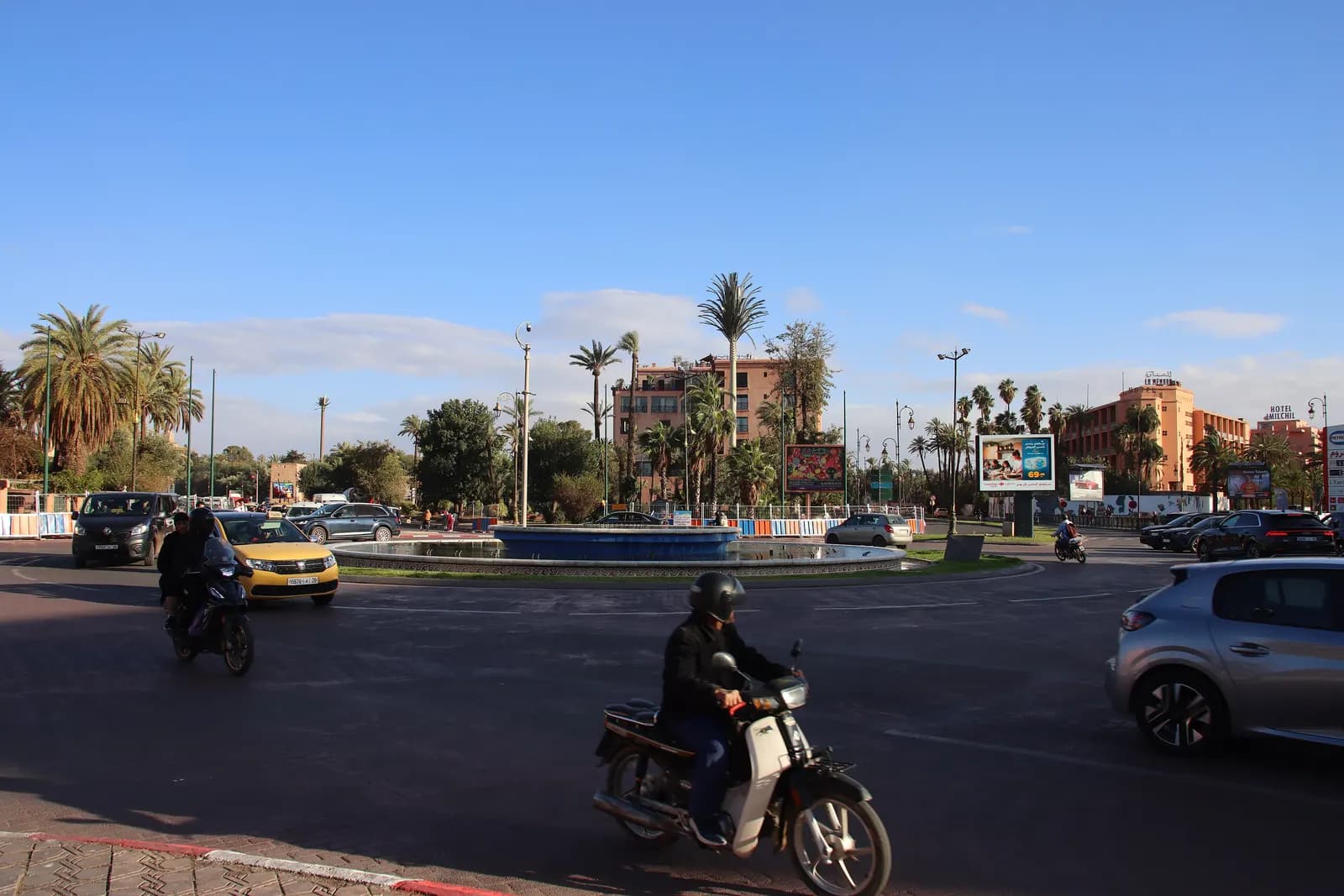 The roundabout at Place de la Liberté in Guéliz, with a fountain, palm trees, hotels behind, a taxi and mopeds on the road