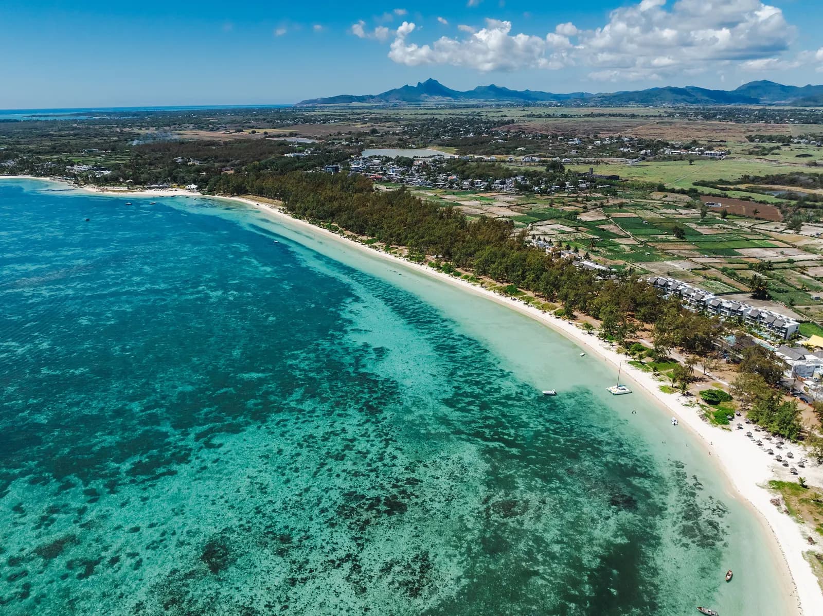 Aerial view of Belle Mare on the east coast, a long white beach between a turquoise lagoon and casuarina trees, with low buildings and fields inland