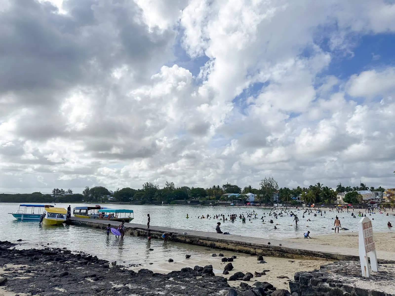 Boats moored at a concrete jetty by the public beach at Blue Bay, with swimmers in the shallow water and houses among trees along the shore