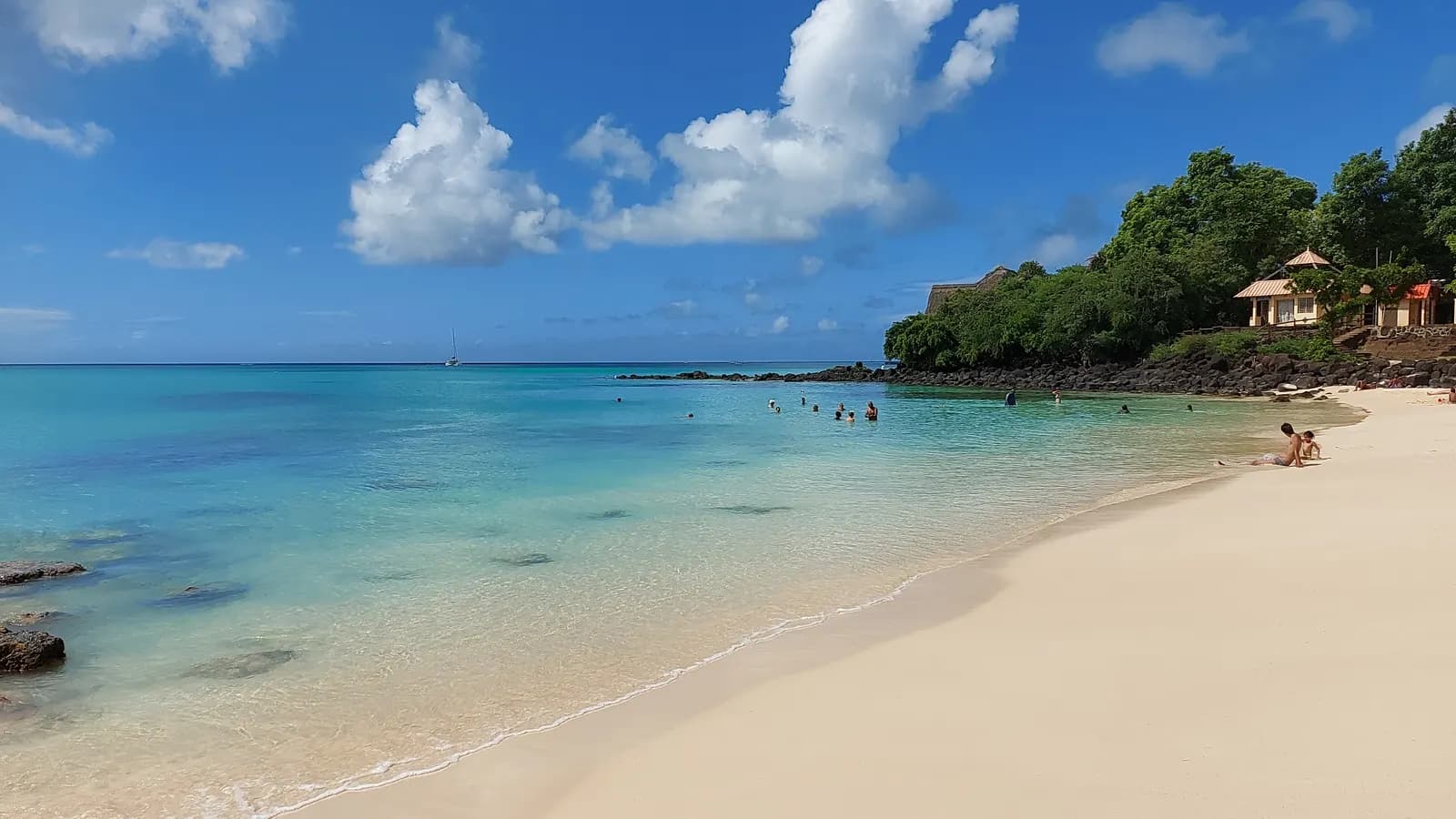 La Cuvette public beach at Grand Baie, with white sand, clear turquoise water, a few swimmers and a small house among trees on the rocky point