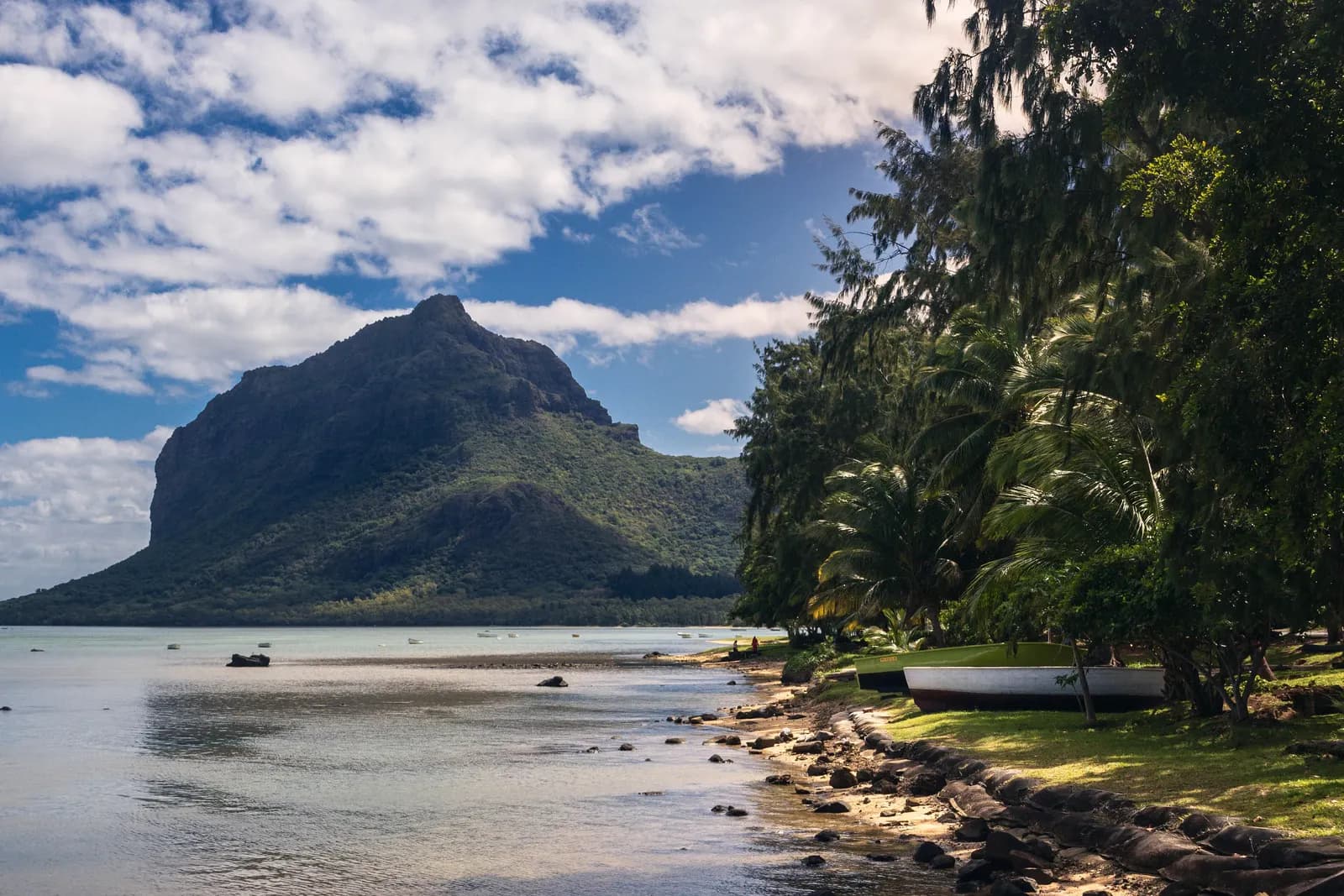 Le Morne Brabant rising above a calm lagoon on the south-west coast of Mauritius, with two boats pulled up on a grassy shore under casuarina and palm trees
