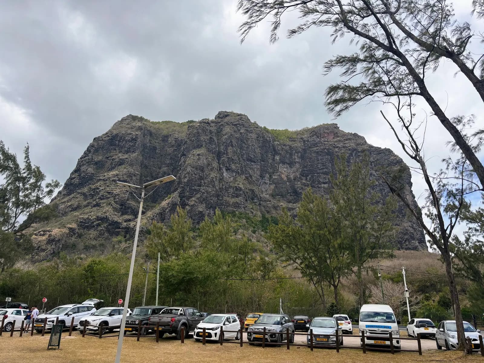 Cars parked in an open car park by the public beach at Le Morne, below the rock face of Le Morne Brabant, under a cloudy sky