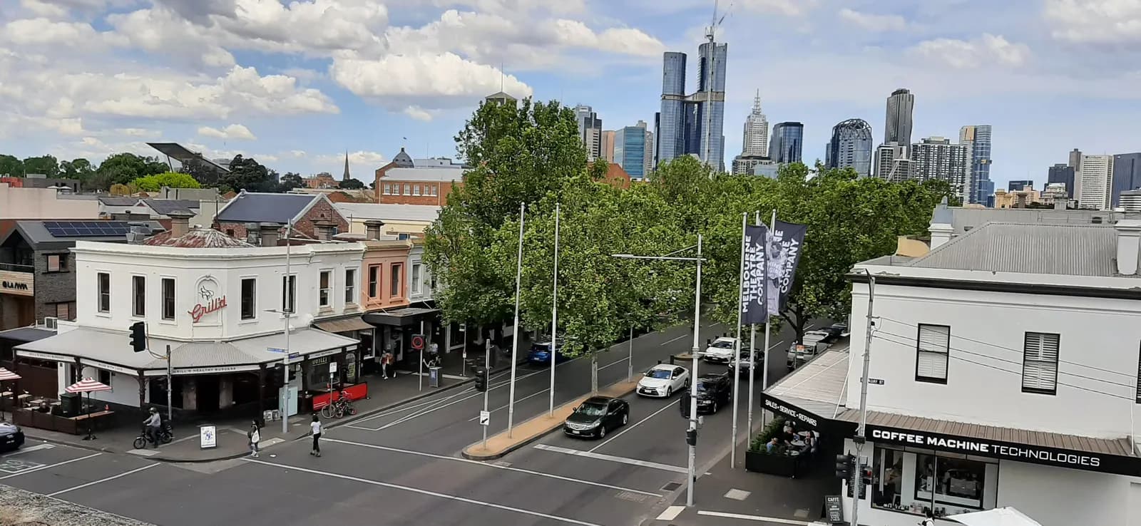 Lygon Street in Carlton, Melbourne, seen from above, with two-storey shopfronts, outdoor tables, a tree-lined median and the city centre towers behind