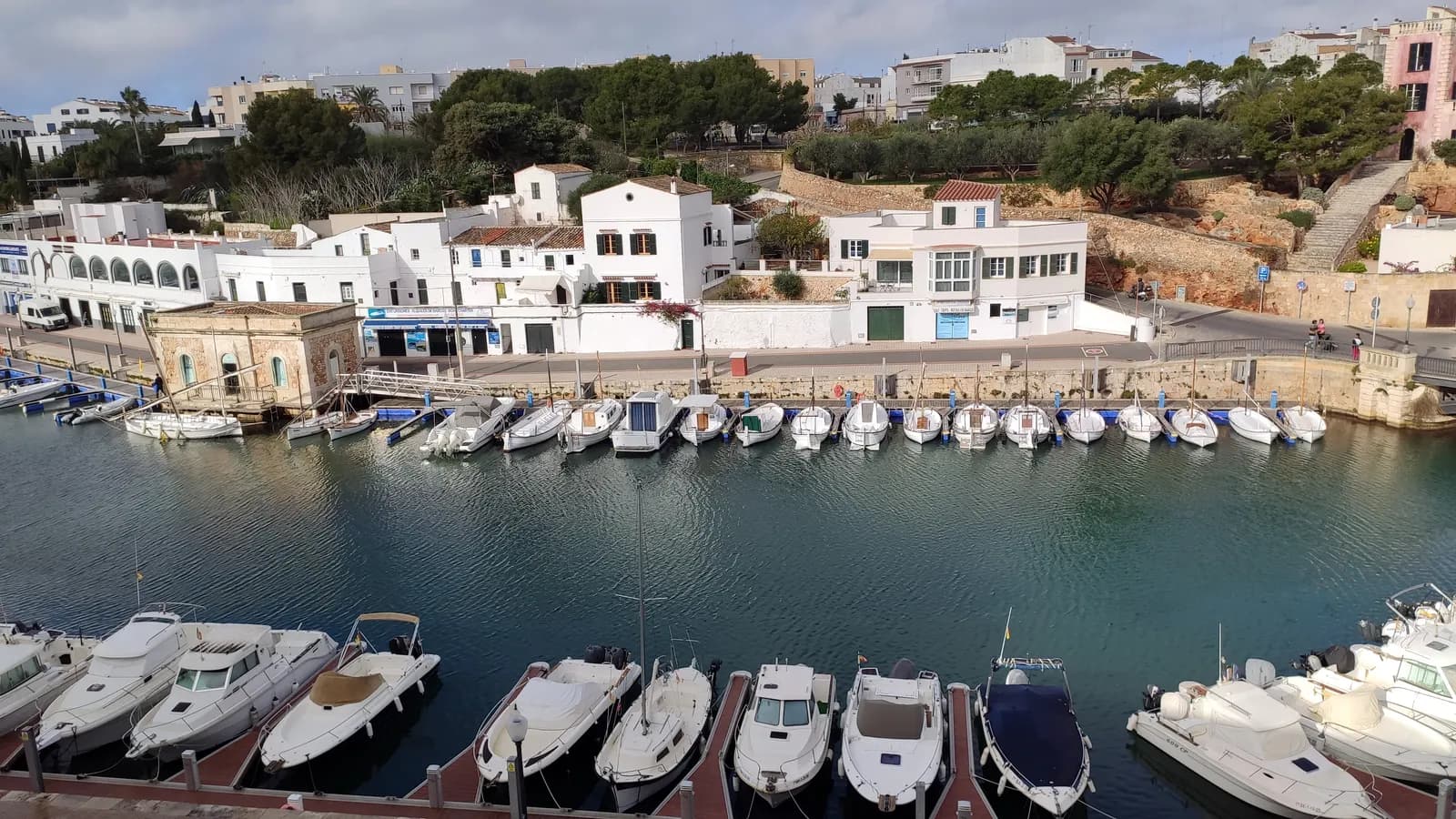 The narrow harbour of Ciutadella with small boats moored along the quay and white houses above it.