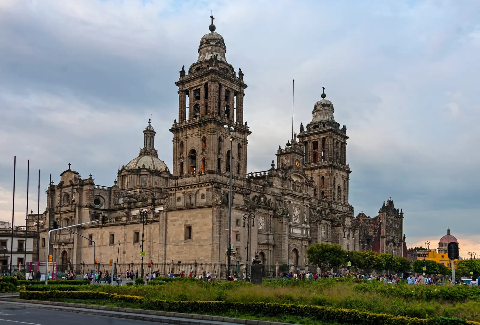 The Metropolitan Cathedral of Mexico City from the corner of the Zócalo, with its stone bell towers and dome above a planted strip and people walking along the railings under a cloudy sky