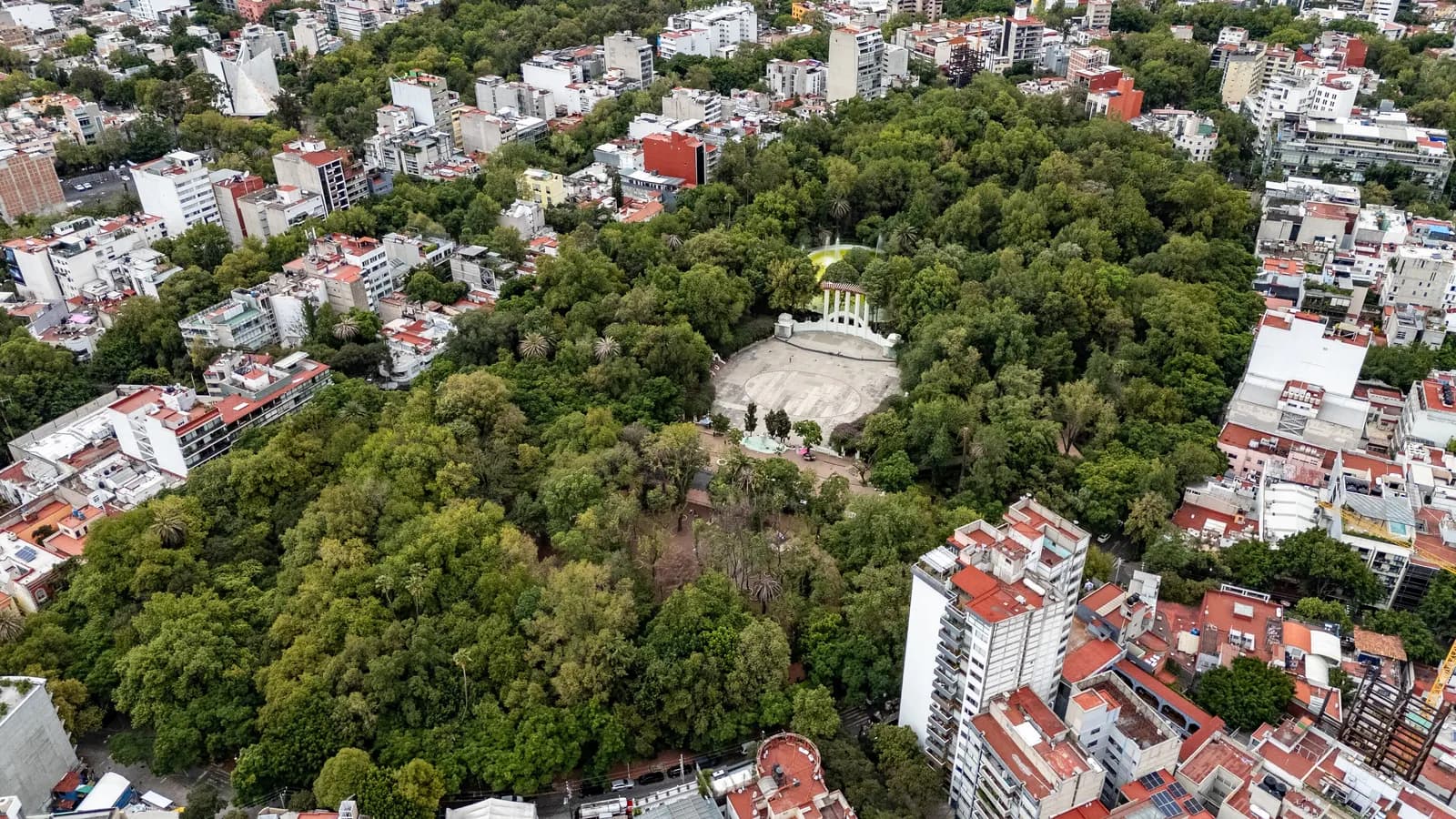 Parque México from the air, a dense canopy of trees around an open-air stage with a pergola, surrounded by the apartment buildings of the Condesa area