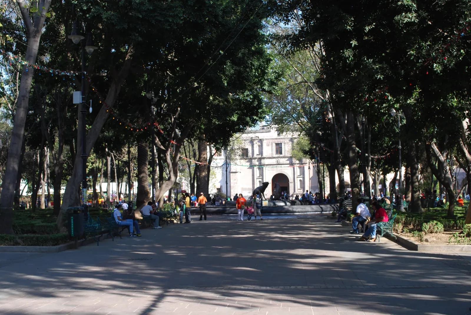 Jardín Centenario in Coyoacán, a paved walk under tall trees with people on benches, a fountain with a coyote statue and the church of San Juan Bautista at the end