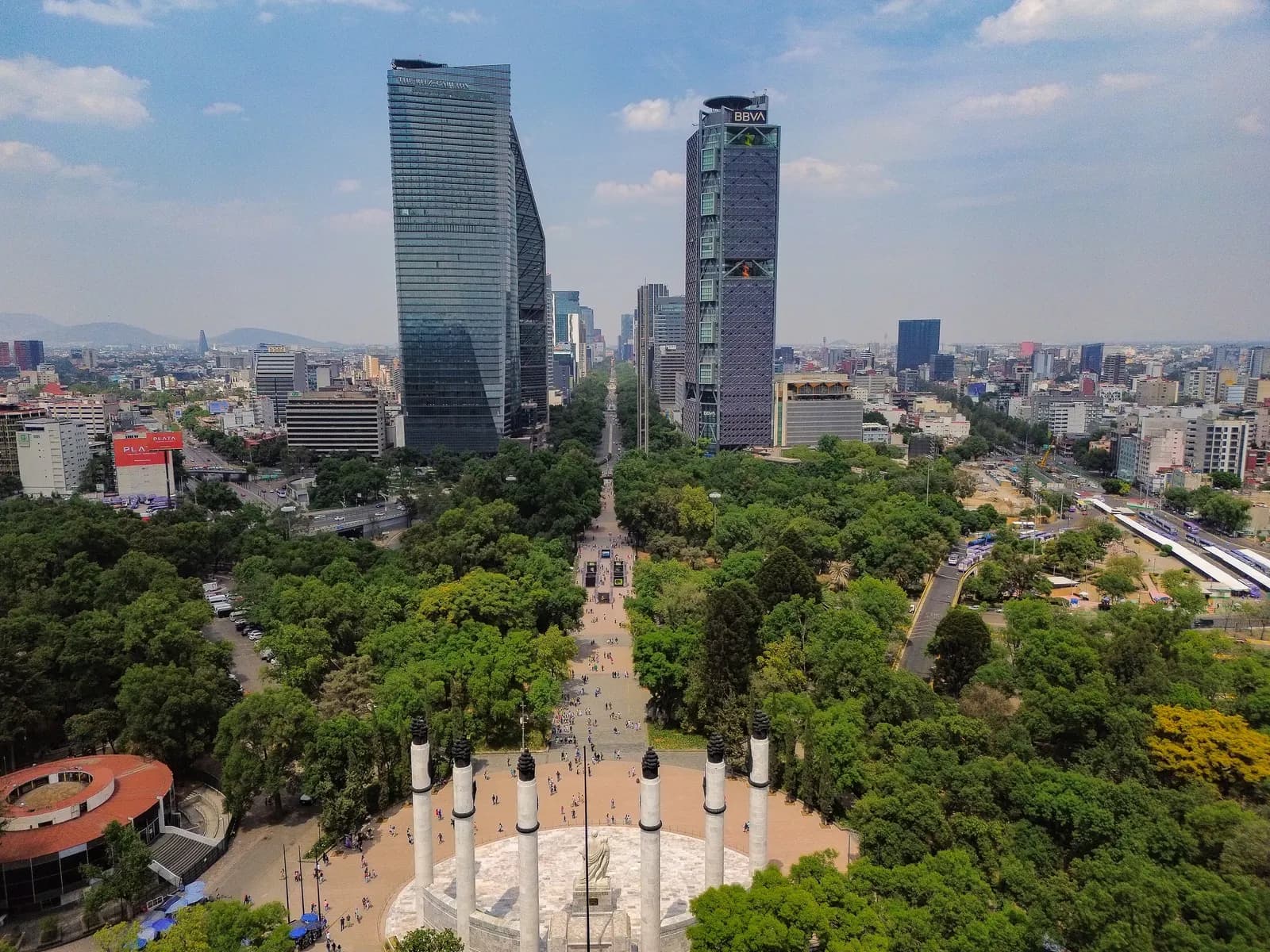 Paseo de la Reforma seen from the air above Chapultepec park, with a semicircle of white columns among the trees, glass office towers on both sides of the avenue and the city stretching to the hills