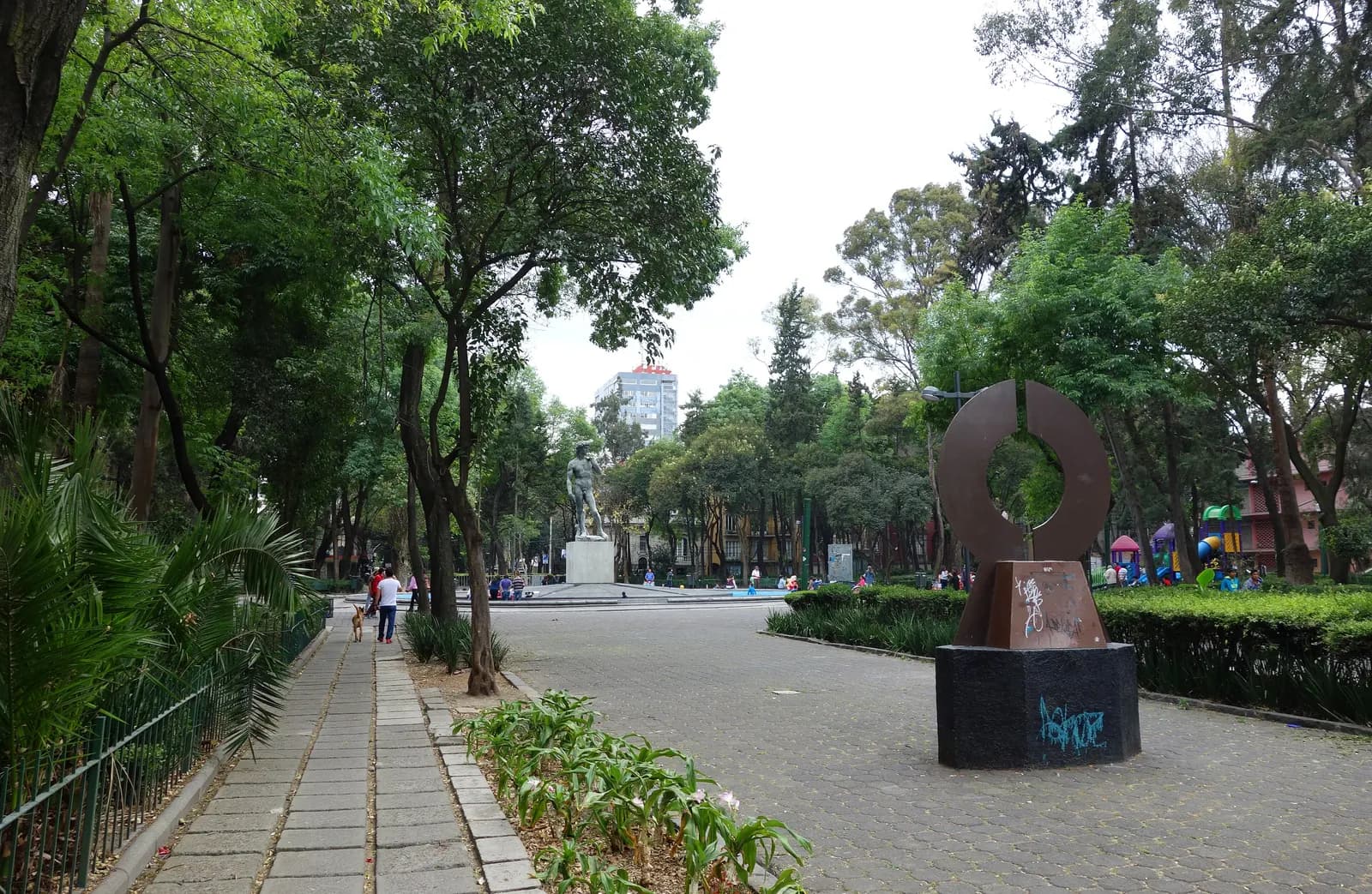 Plaza Río de Janeiro in Colonia Roma, a paved square shaded by tall trees, with a copy of Michelangelo's David on a plinth, a round metal sculpture and people walking in the square