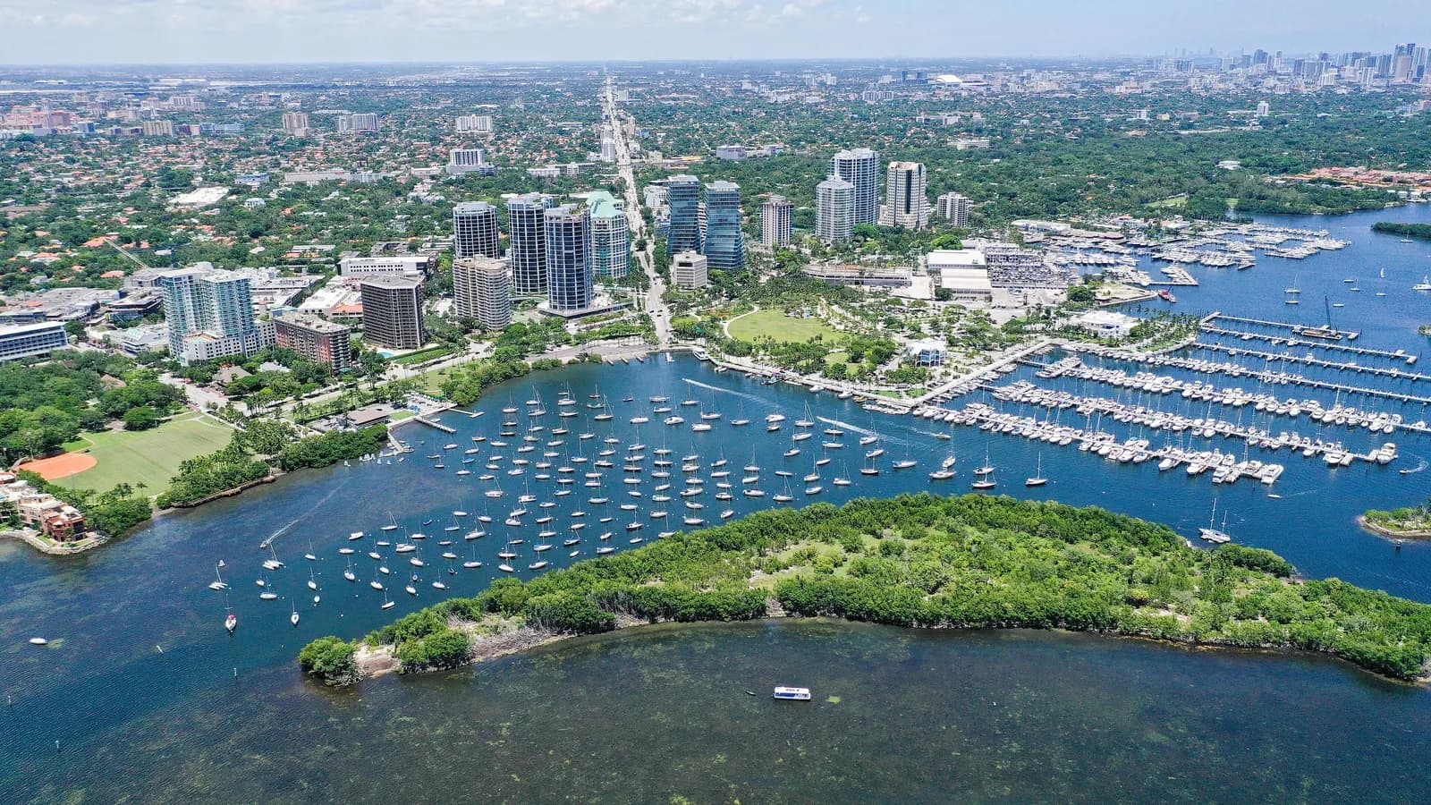 Aerial view of Coconut Grove, with towers among dense trees, sailboats moored in the bay and marinas along the shore, and the Miami skyline far in the distance