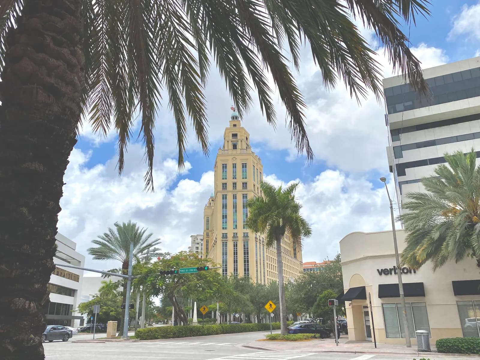 A tall cream-coloured tower with a domed top rising behind palm trees at a street corner in Coral Gables, with shops and offices on either side
