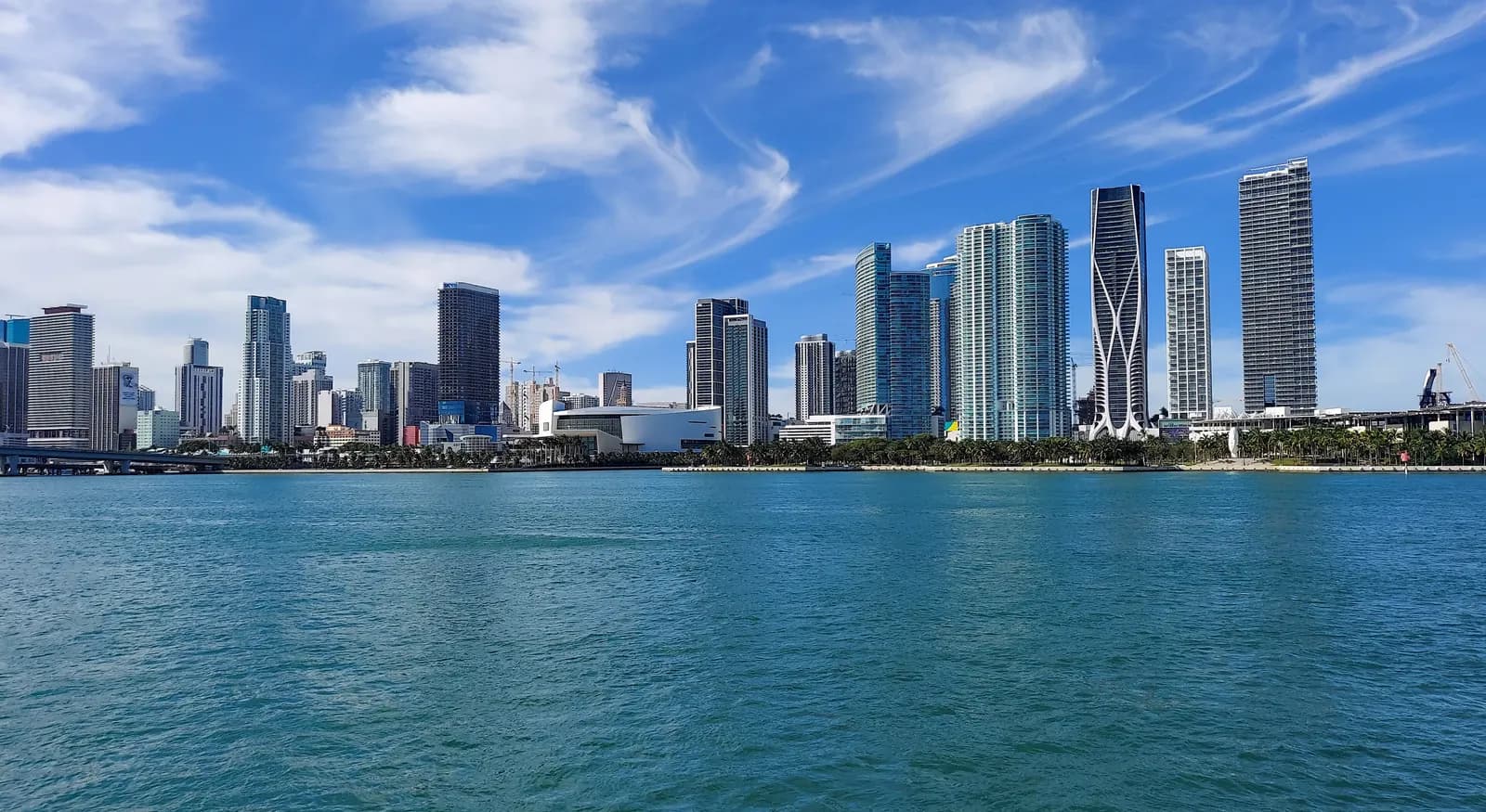 Downtown Miami towers and a low white arena along the palm-lined waterfront, seen across the blue water of Biscayne Bay under a blue sky with thin clouds