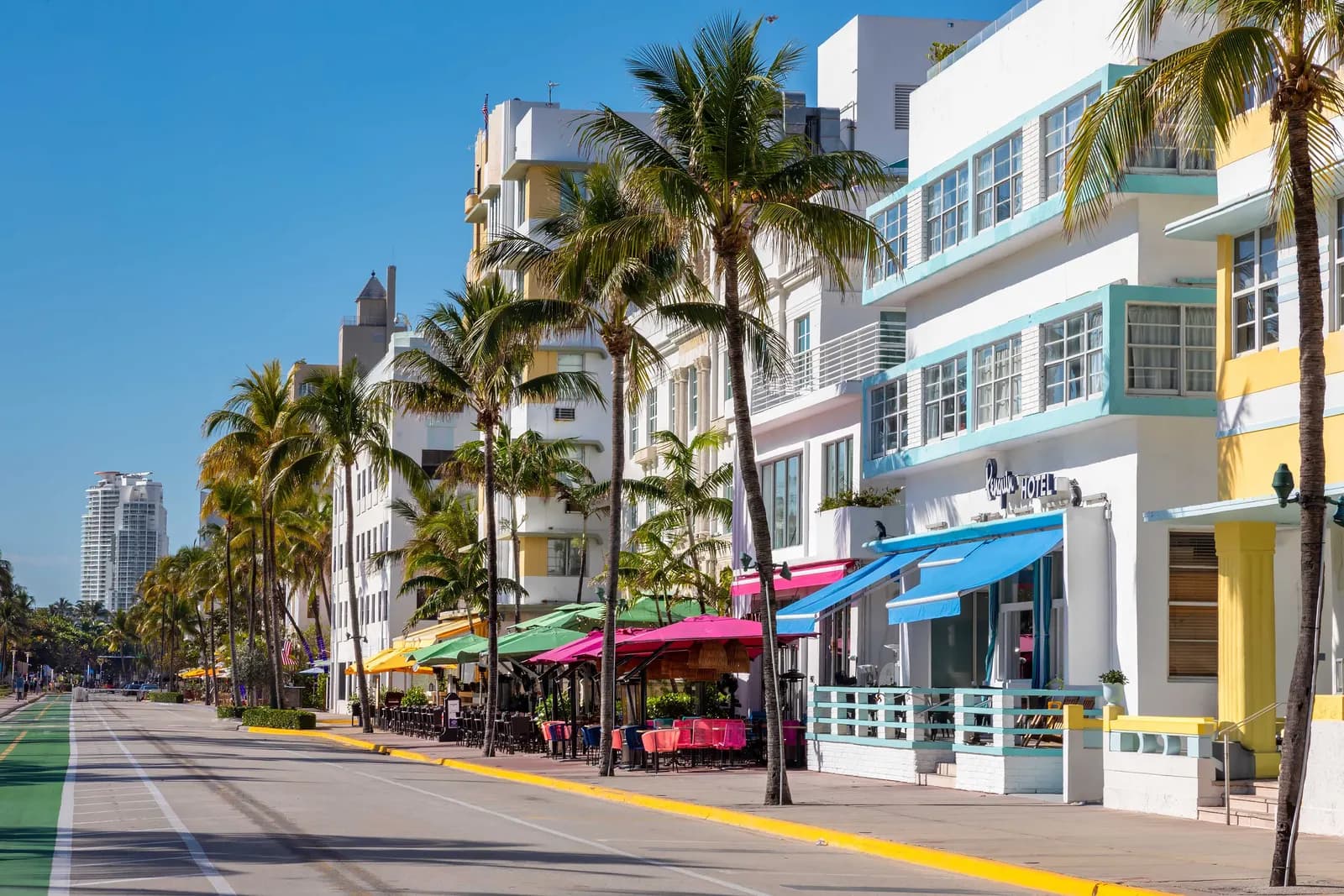 Pastel art deco hotels with blue and yellow trim and café umbrellas behind a row of palm trees on Ocean Drive in South Beach, under a clear blue sky