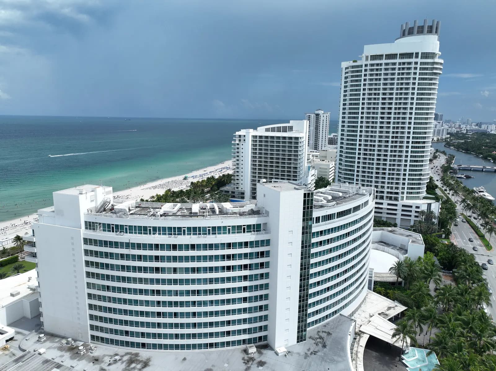 The curved white Fontainebleau hotel and other hotel towers along the beach at Mid-Beach seen from the air, with a palm-lined road and a canal with boats on the right and a grey sky over the sea