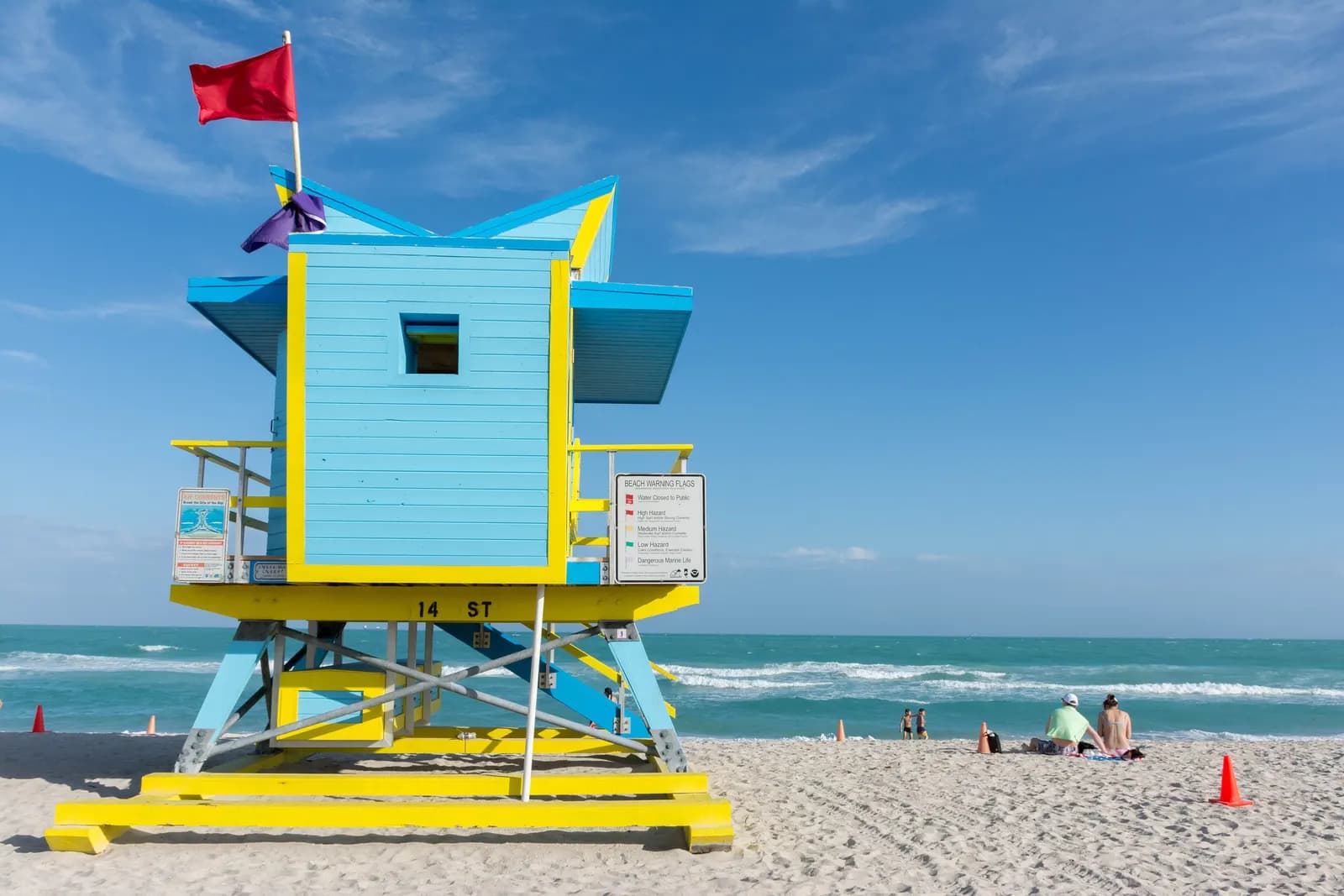A light blue and yellow lifeguard tower marked 14 ST on the white sand of South Beach, with a red flag flying, the turquoise sea behind and a few people on the beach